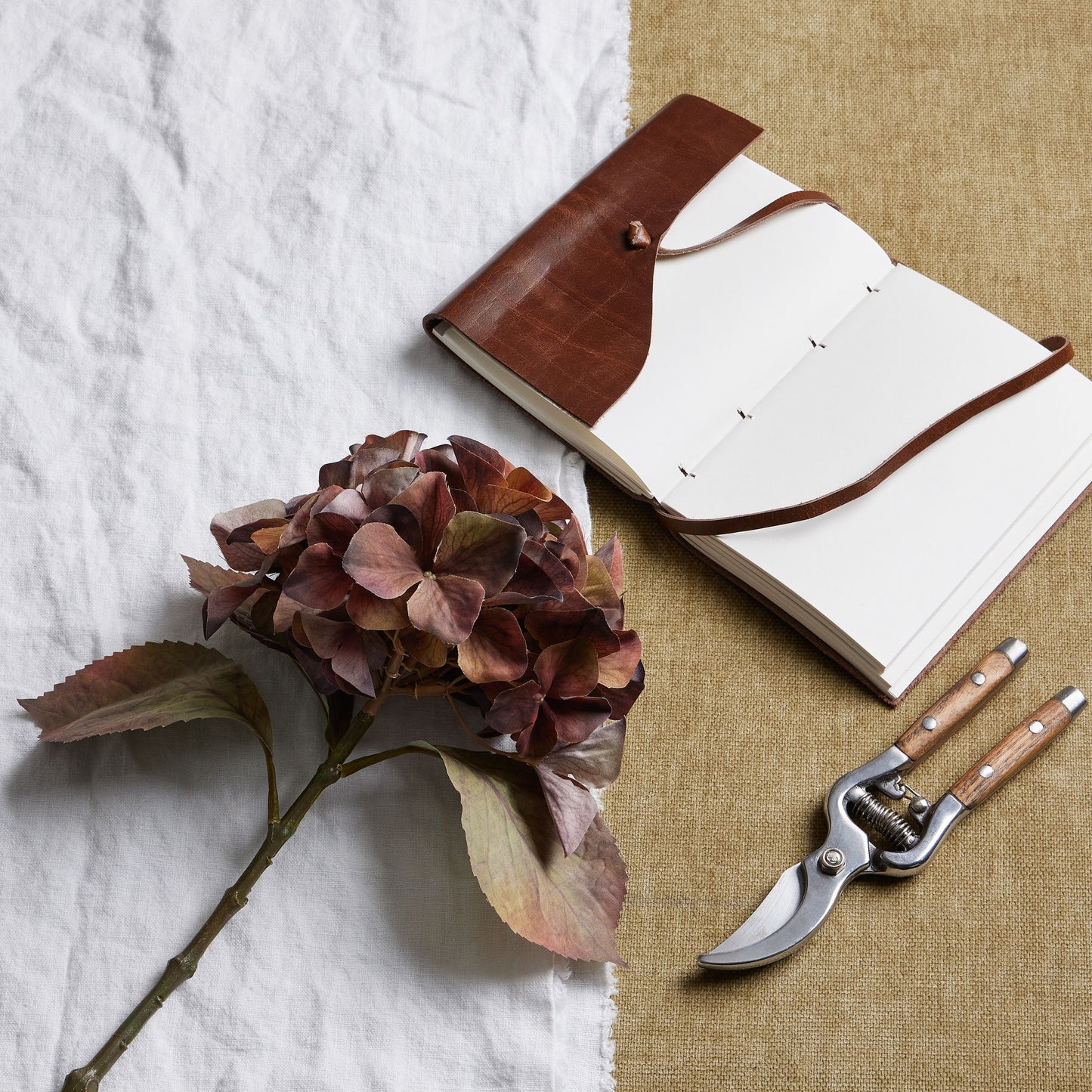 Single hydrangea stem in deep brown, amber, and russet hues, paired with a leather notebook and pruning shears on textured fabric.