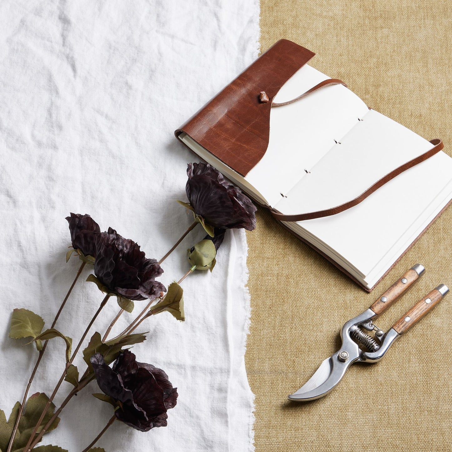 Tall black poppy stem with realistic petals, placed beside a brown leather notebook and pruning shears on textured fabric.