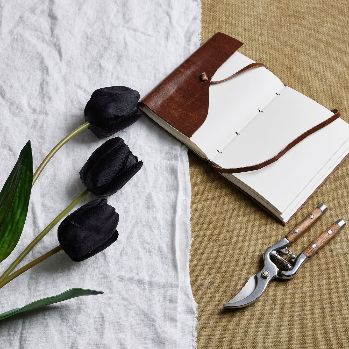 Tall black triple tulip stems arranged with a leather-bound notebook and garden shears on textured fabric, exuding elegance.