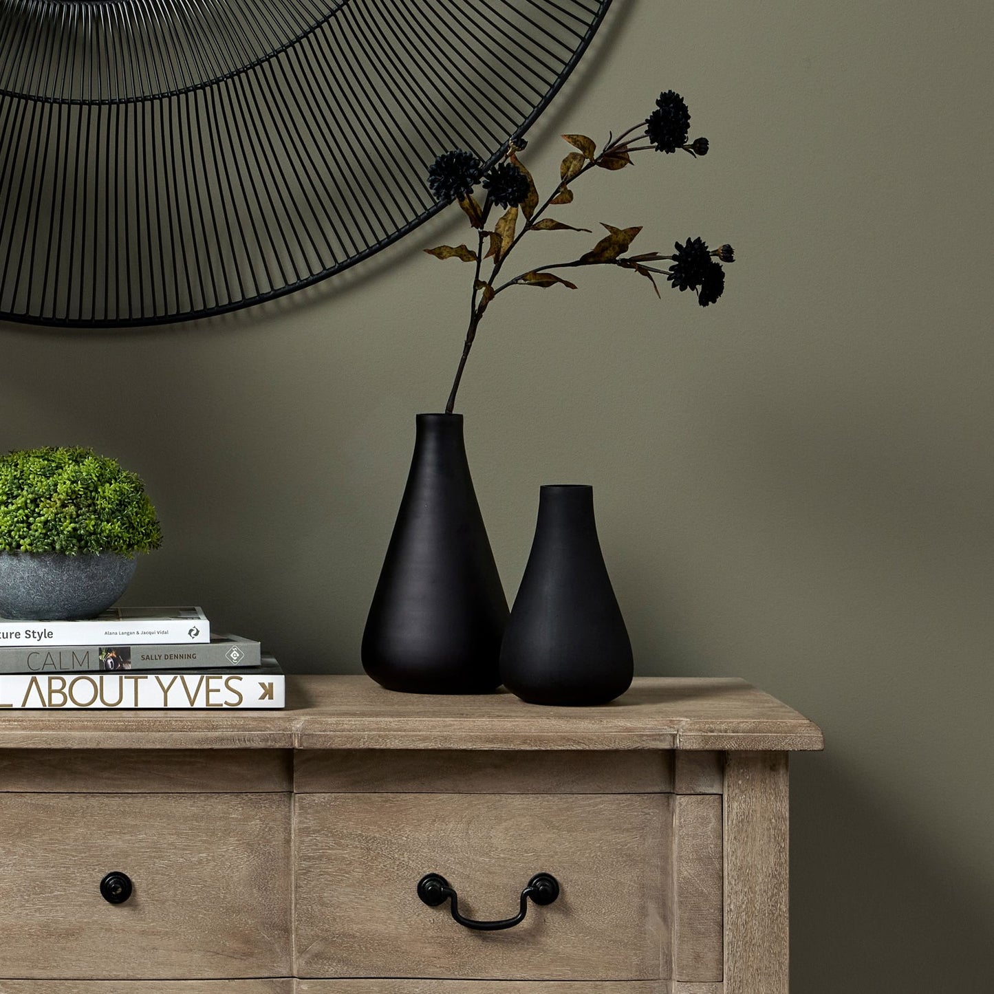 Sleek black tapered vase on a wooden table, accented with dark stems, enhancing a modern interior against a green wall.