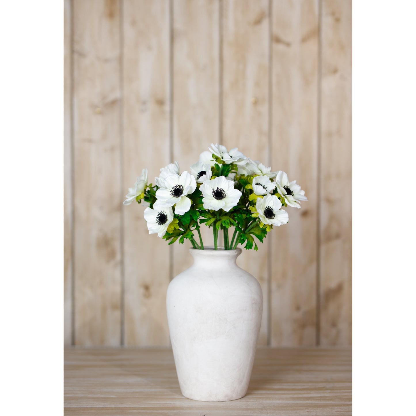 White anemone spray in a smooth, white vase, showcasing delicate petals and dark centers against lush green foliage.
