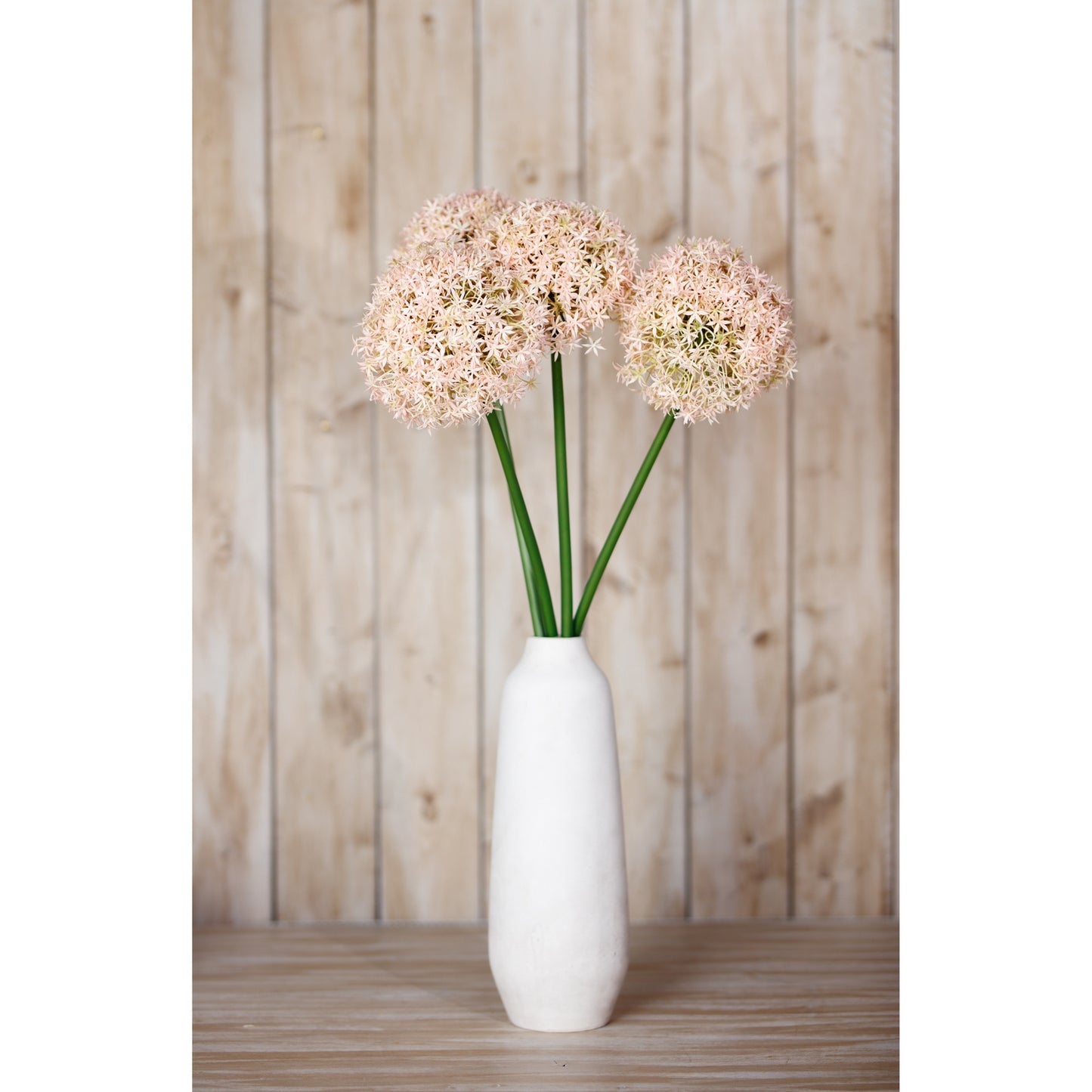 Pink faux allium stem with delicate spherical pink and ivory blooms, displayed in a white vase against a rustic wooden backdrop.