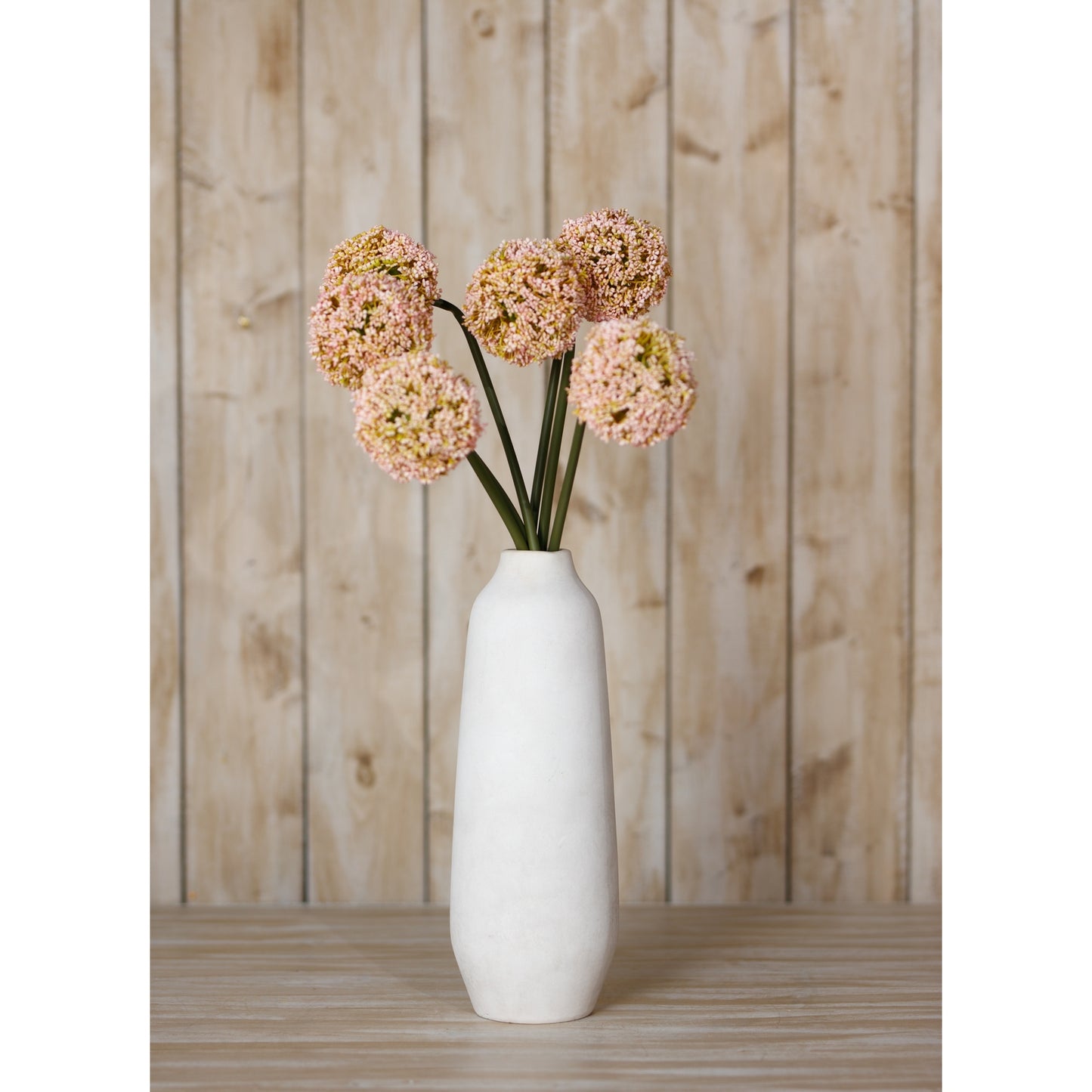 Pink allium stems arranged in a white vase, showcasing delicate floral clusters with vibrant pink hues against a rustic wooden backdrop.
