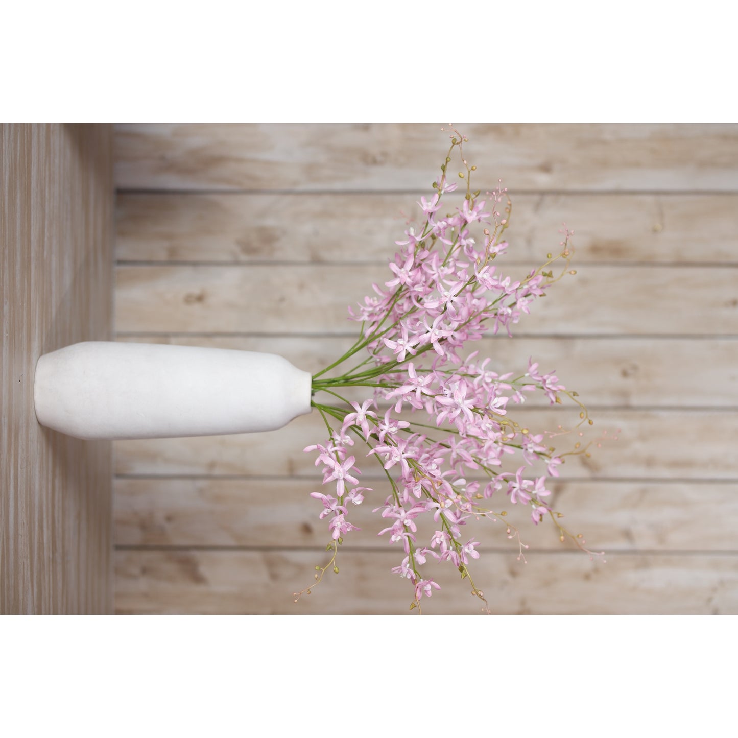 Delicate lilac cymbidium spray in a white vase, showcasing soft lavender petals and graceful cascading stems against a rustic wooden backdrop.