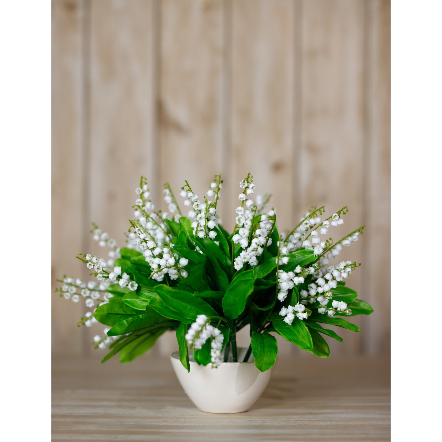 Bunch of artificial Lily of the Valley flowers with white blooms and lush green leaves in a cream-colored vase, set against a wooden backdrop.