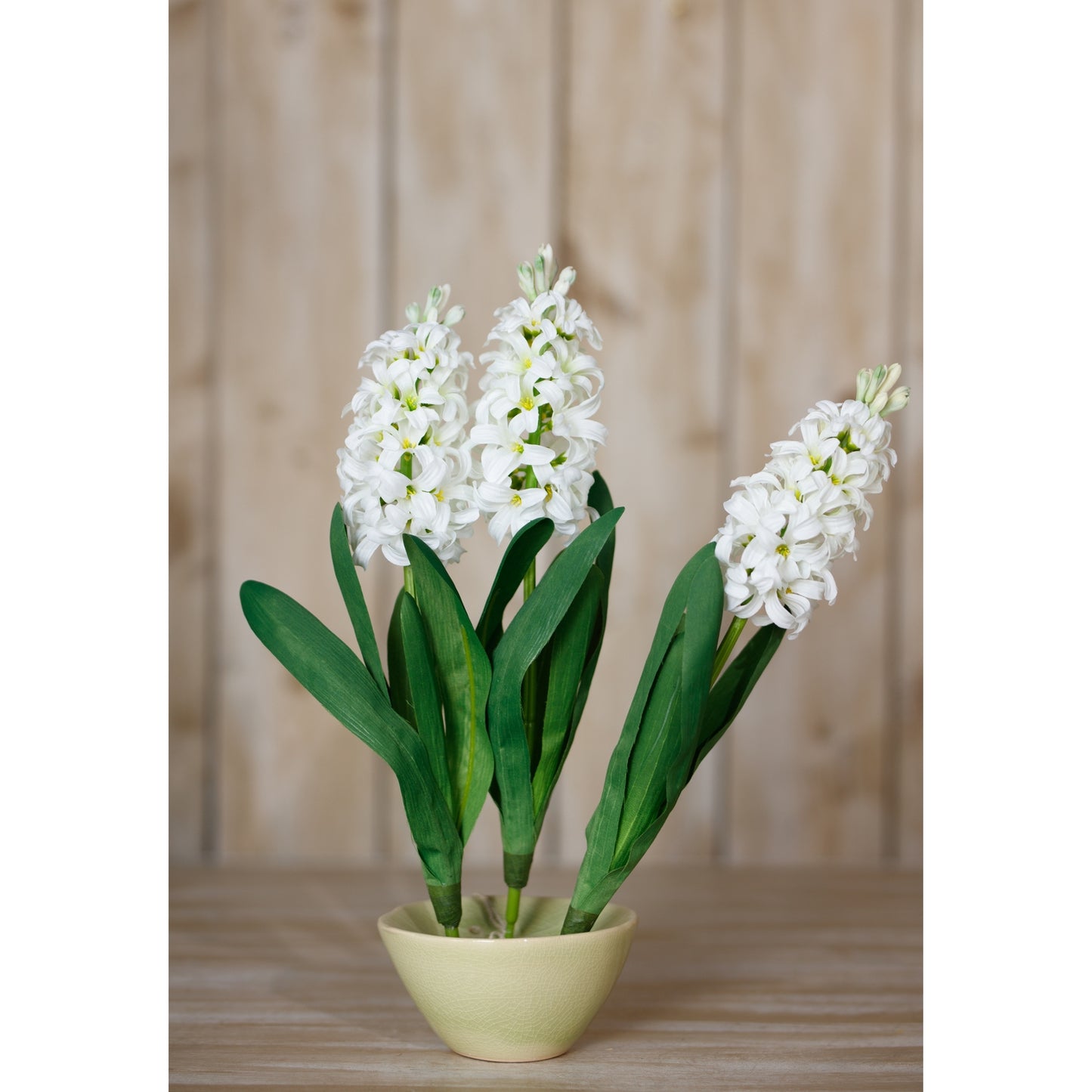 Elegant white hyacinth flowers with lush green leaves in a pale yellow bowl, set against a soft, neutral background.