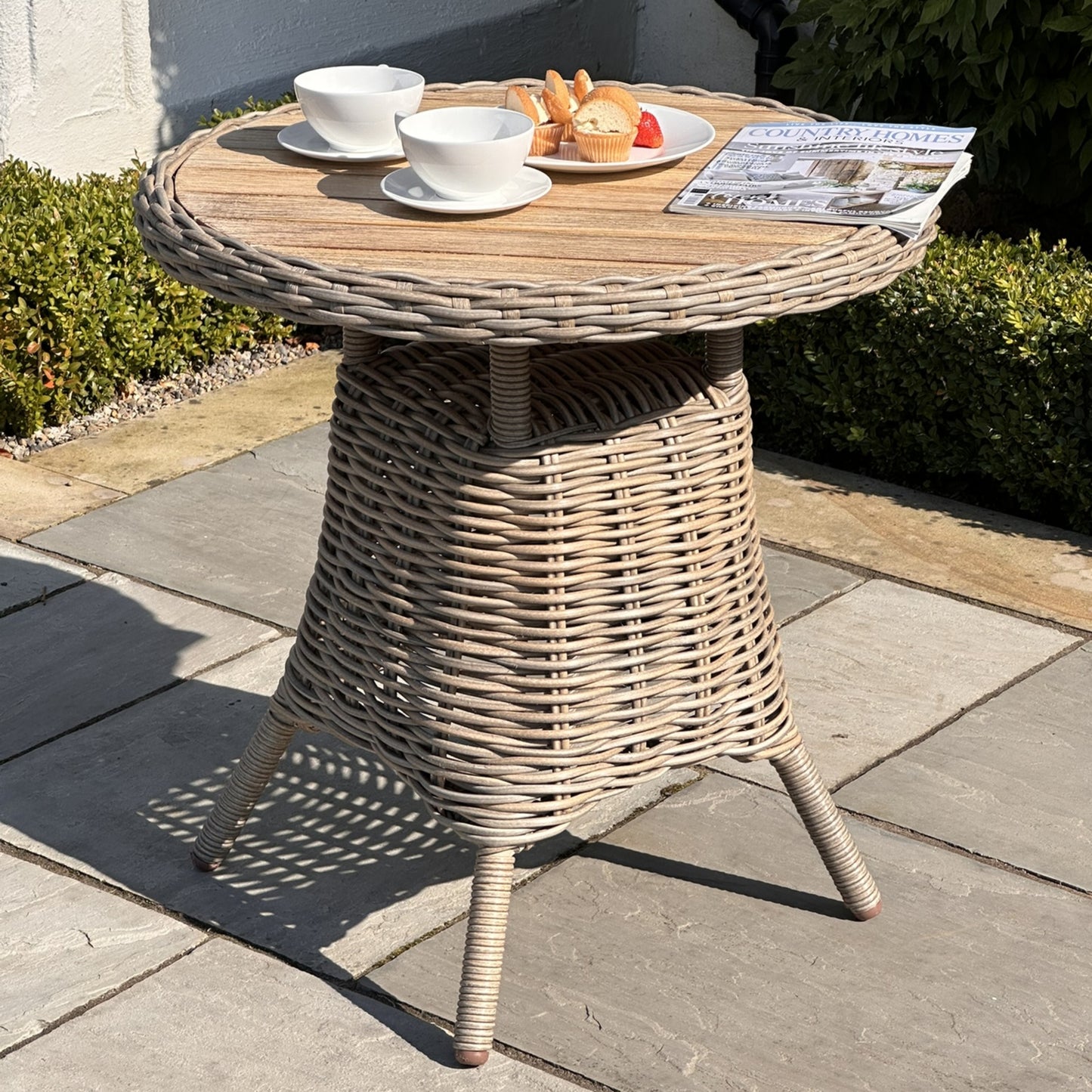 Elegant outdoor bistro table with a natural wood top, woven base, and light gray finish, set with coffee cups and pastries.