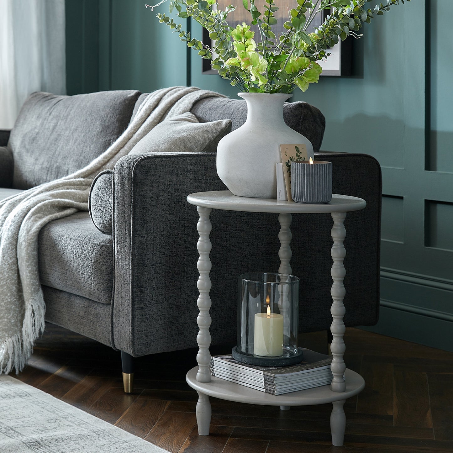 Elegant round side table in soft gray, featuring turned legs, a stacked book base, and a vase of greenery, complementing modern decor.