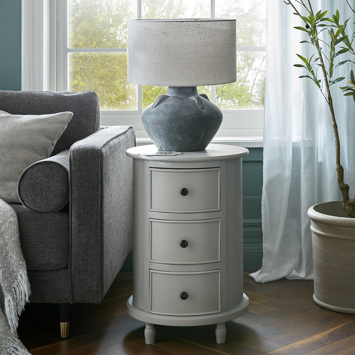 Sophisticated round side table in light gray, featuring three drawers and topped with a textured, stone-like lamp, beside a cozy couch.