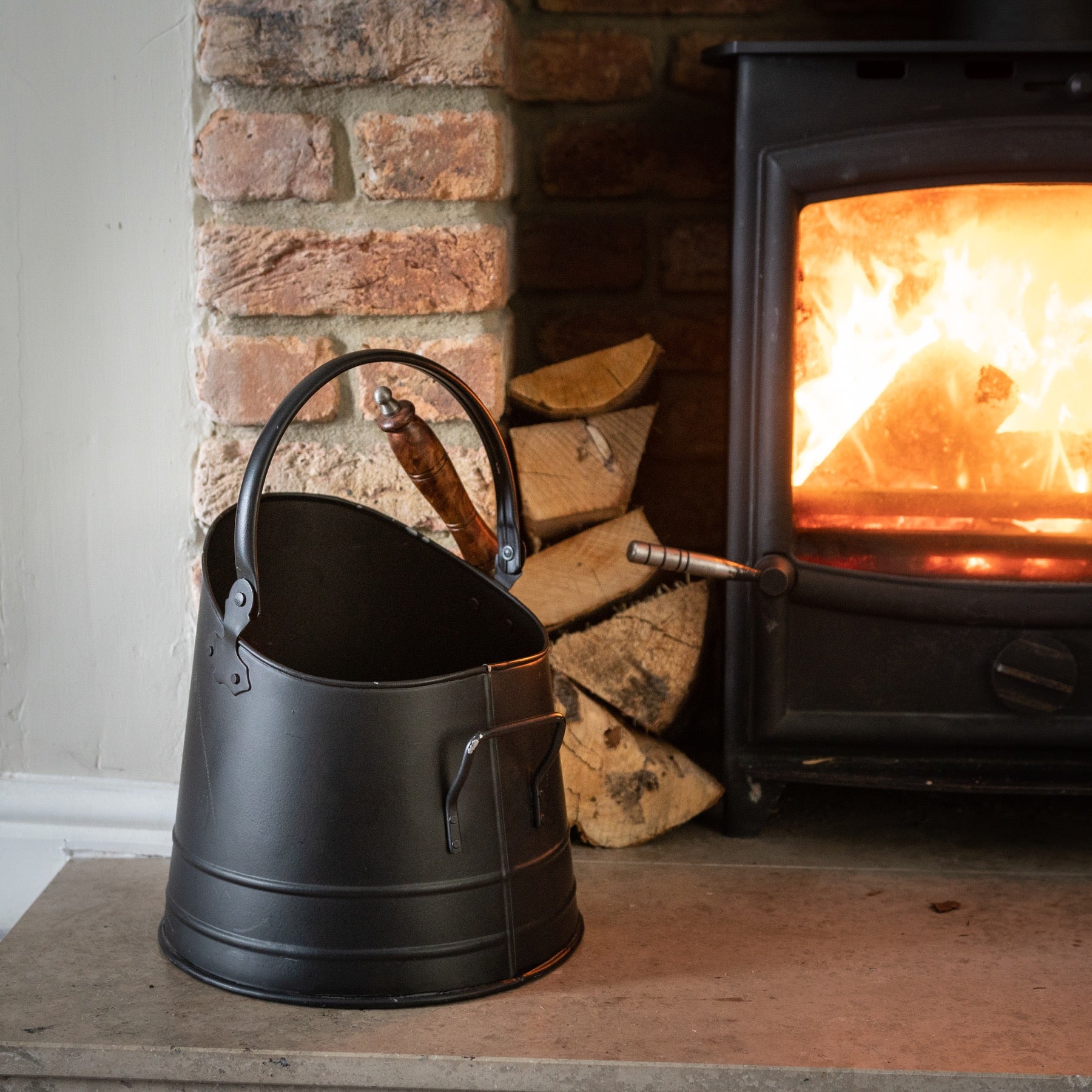 Black coal bucket made of durable steel with a smooth teak handle, resting near a fireplace and stacked firewood.