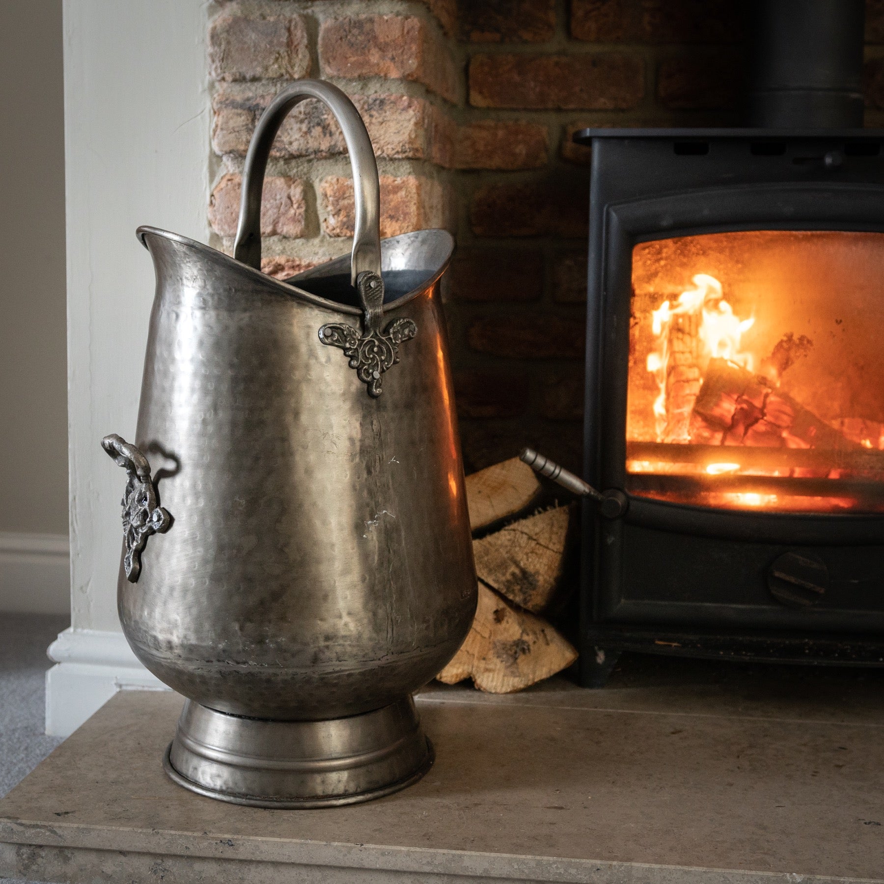 Antique pewter coal bucket with intricate leaf motifs, ornate handle, and a hammered finish, set beside a glowing fireplace.
