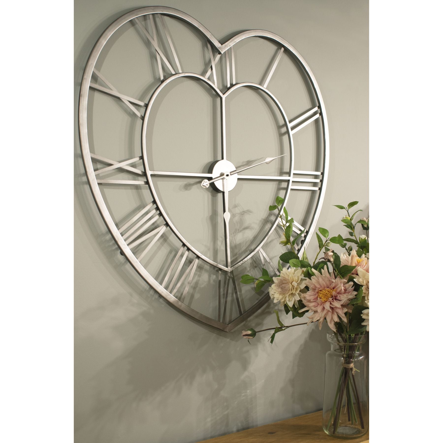 Silver heart-shaped wall clock with Roman numerals and classic hands, featuring a minimalist design against a soft green wall.