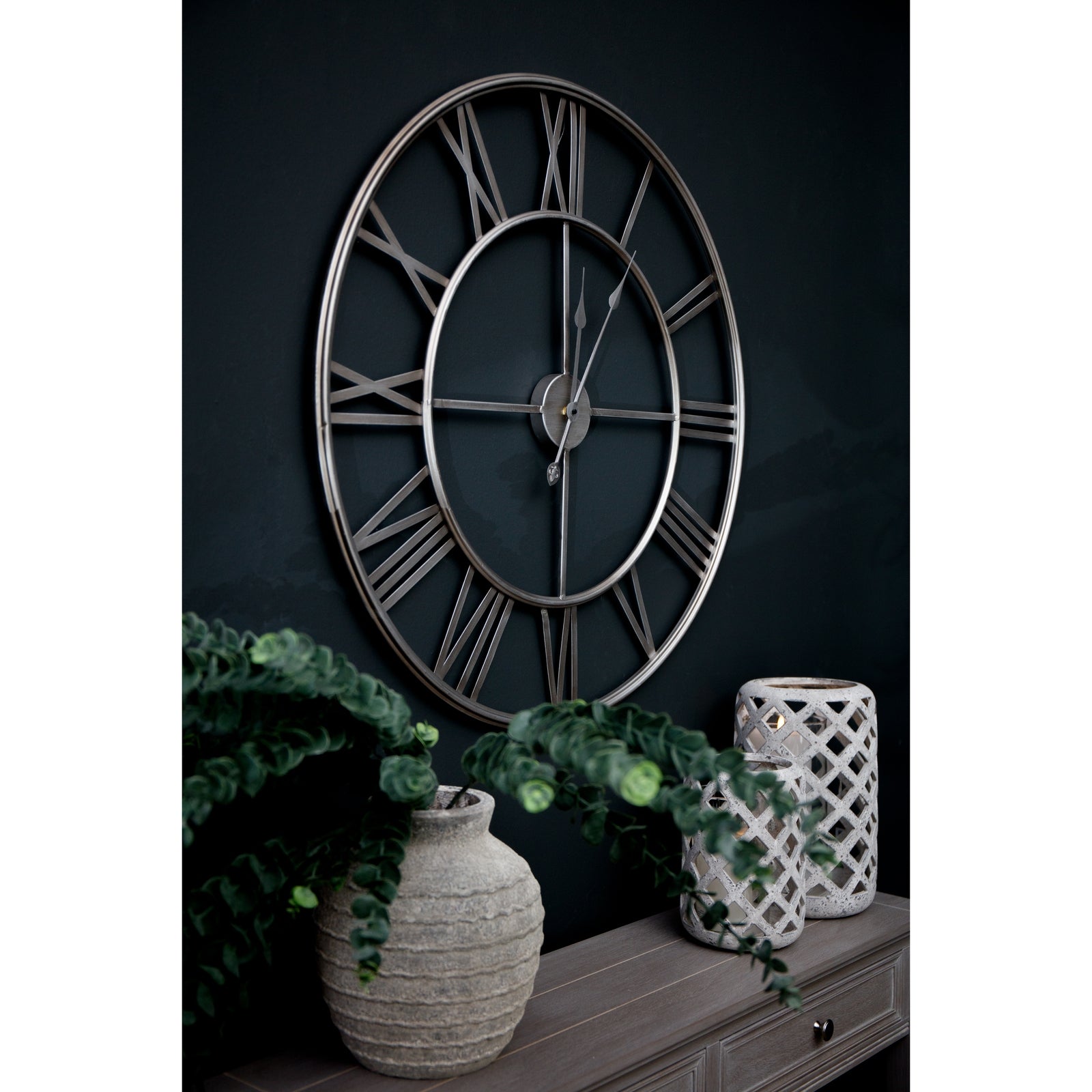 Large silver foil wall clock with intricate metalwork and Roman numerals, set against a dark wall, flanked by decorative vases.