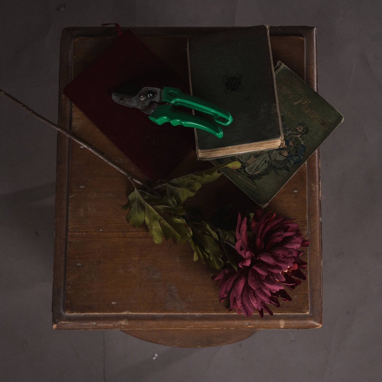 Deep red artificial spider chrysanthemum with intricate petals, resting alongside vintage books and green foliage on a wooden table.