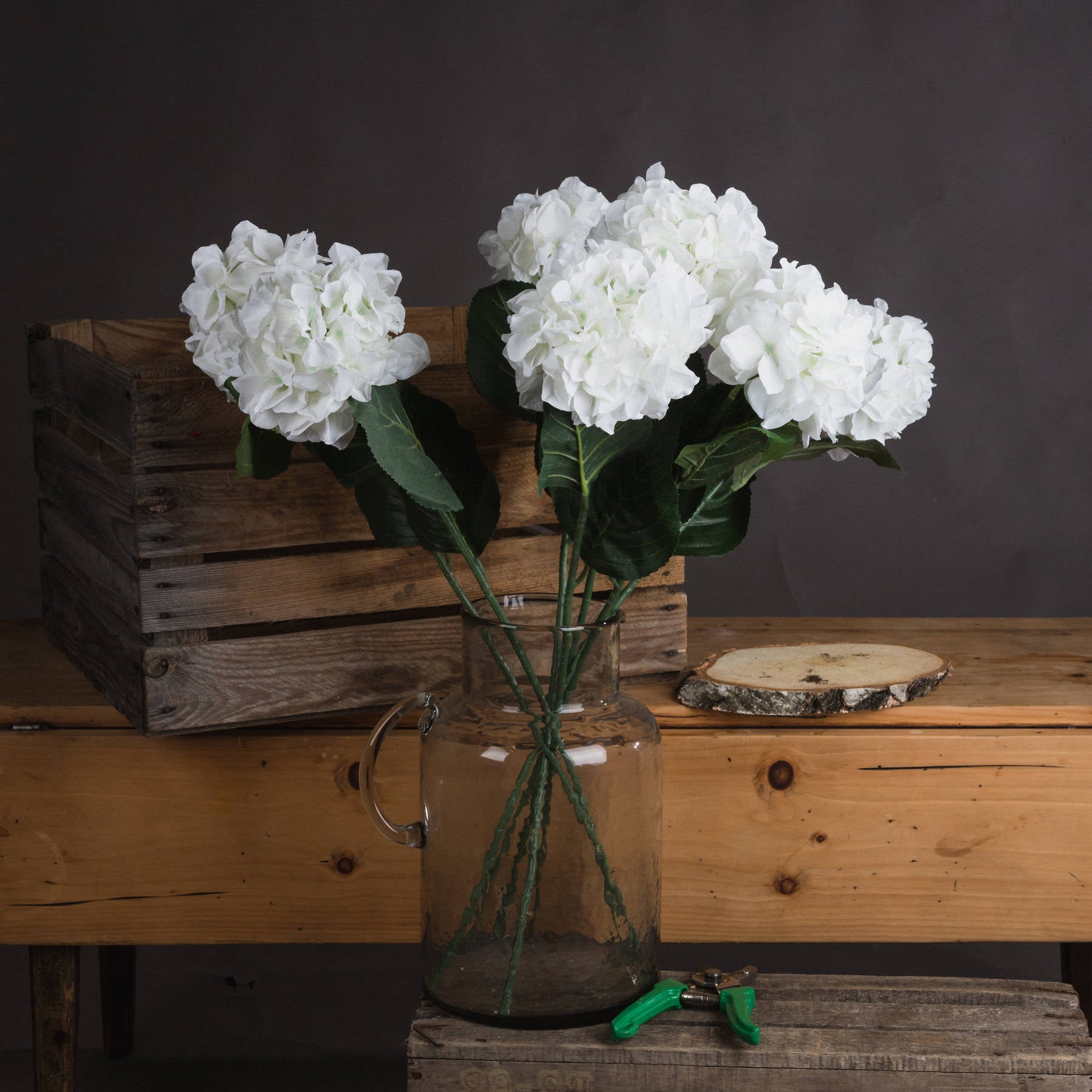 Single stem white hydrangea in a clear glass vase, surrounded by green leaves, on a wooden table with rustic crates.