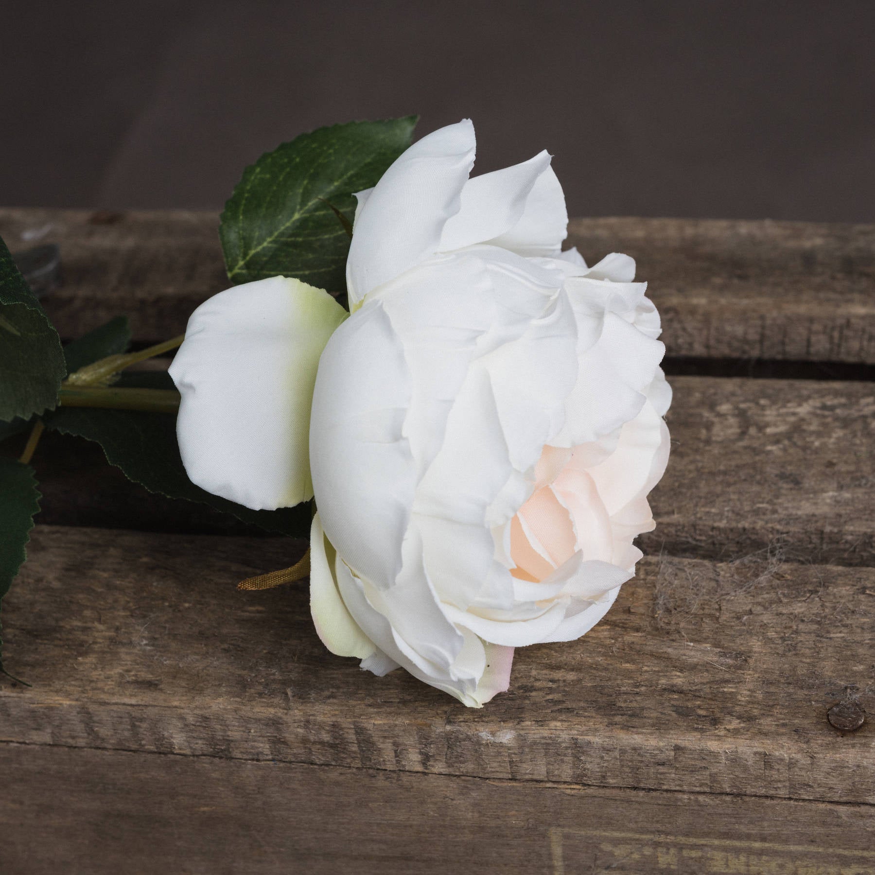 Single stem artificial blush garden rose with soft white petals and subtle peach center, complemented by green leaves, resting on rustic wood.