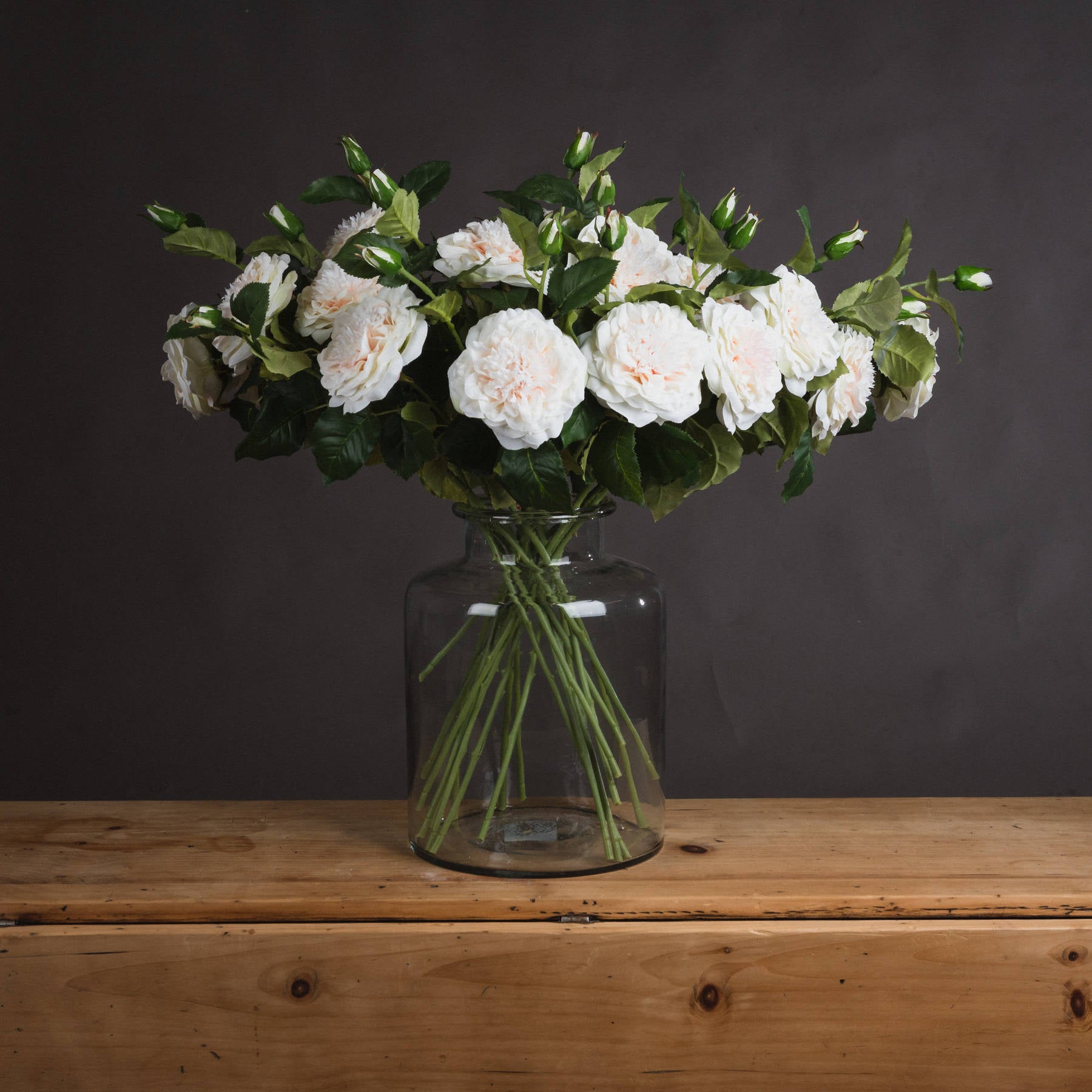 Luxurious artificial white garden roses in a glass vase, showcasing delicate blooms and lush greenery against a dark background.