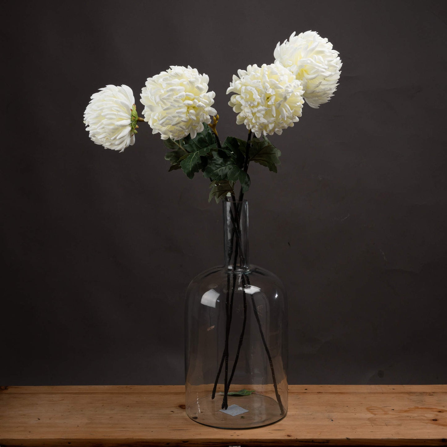 Large white artificial chrysanthemums in a clear glass vase, resting on a wooden surface against a dark background.