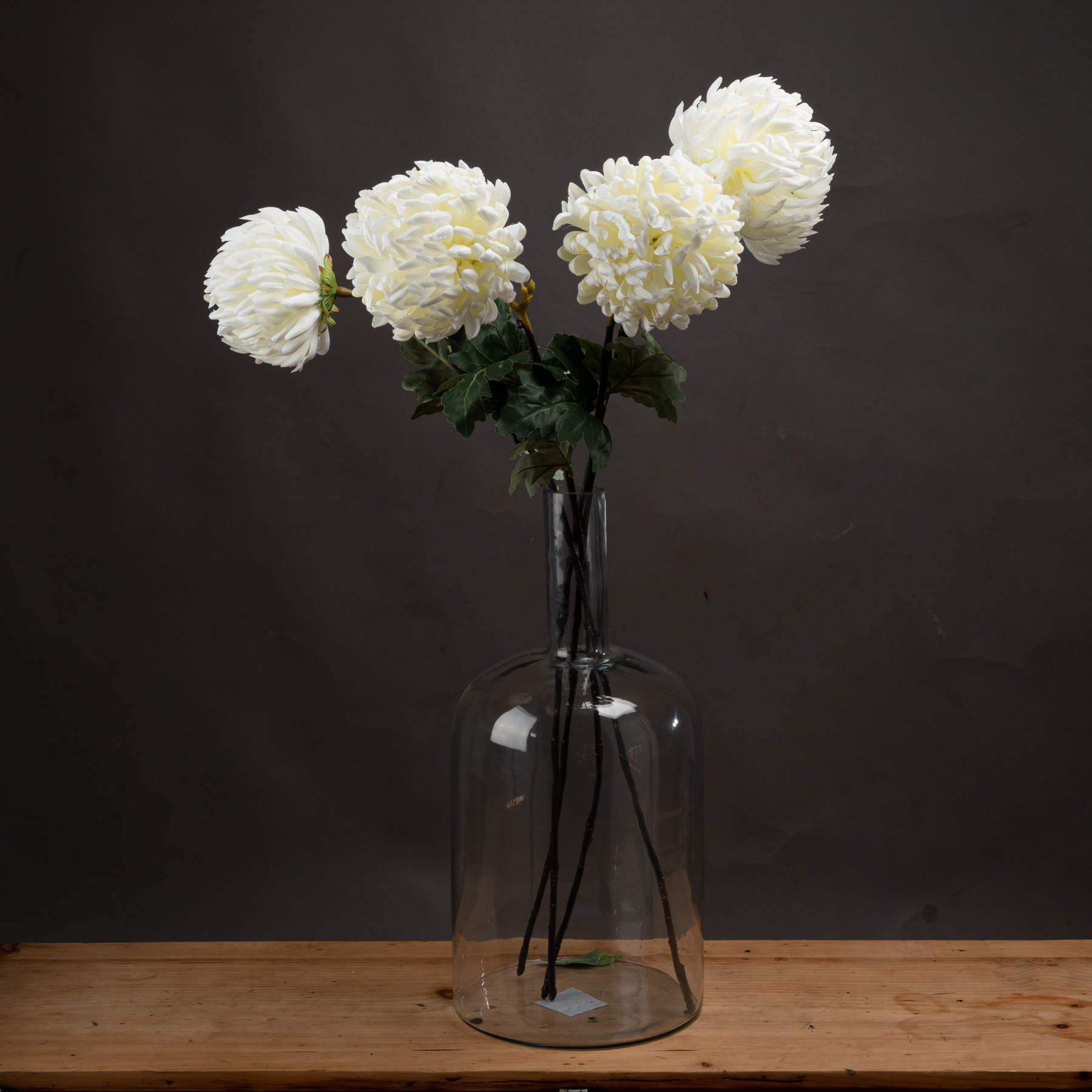 Large white artificial chrysanthemums in a clear glass vase, resting on a wooden surface against a dark background.
