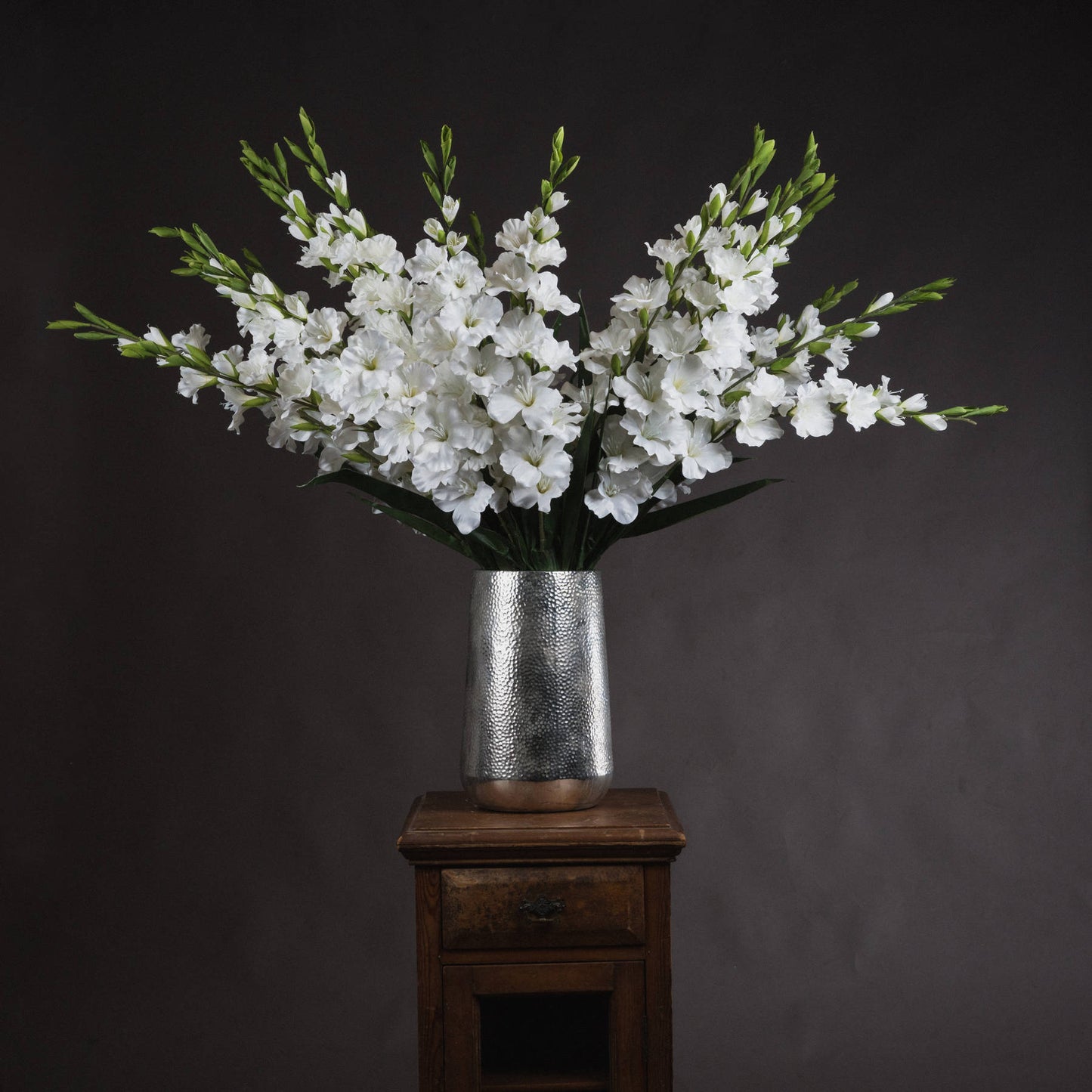 Lifelike white gladioli elegantly arranged in a silver textured vase, showcasing pristine florets and lush green leaves.