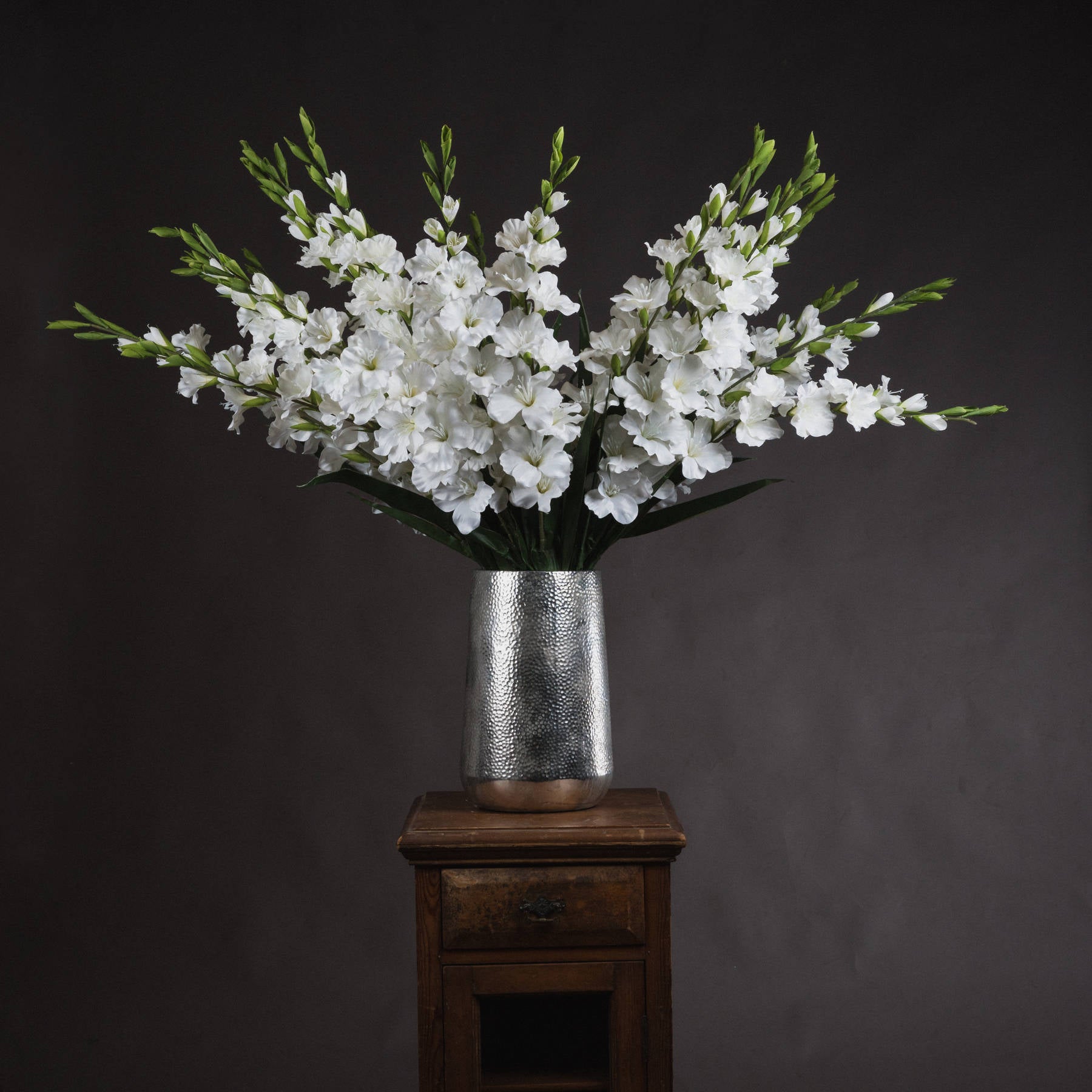 Lifelike white gladioli elegantly arranged in a silver textured vase, showcasing pristine florets and lush green leaves.