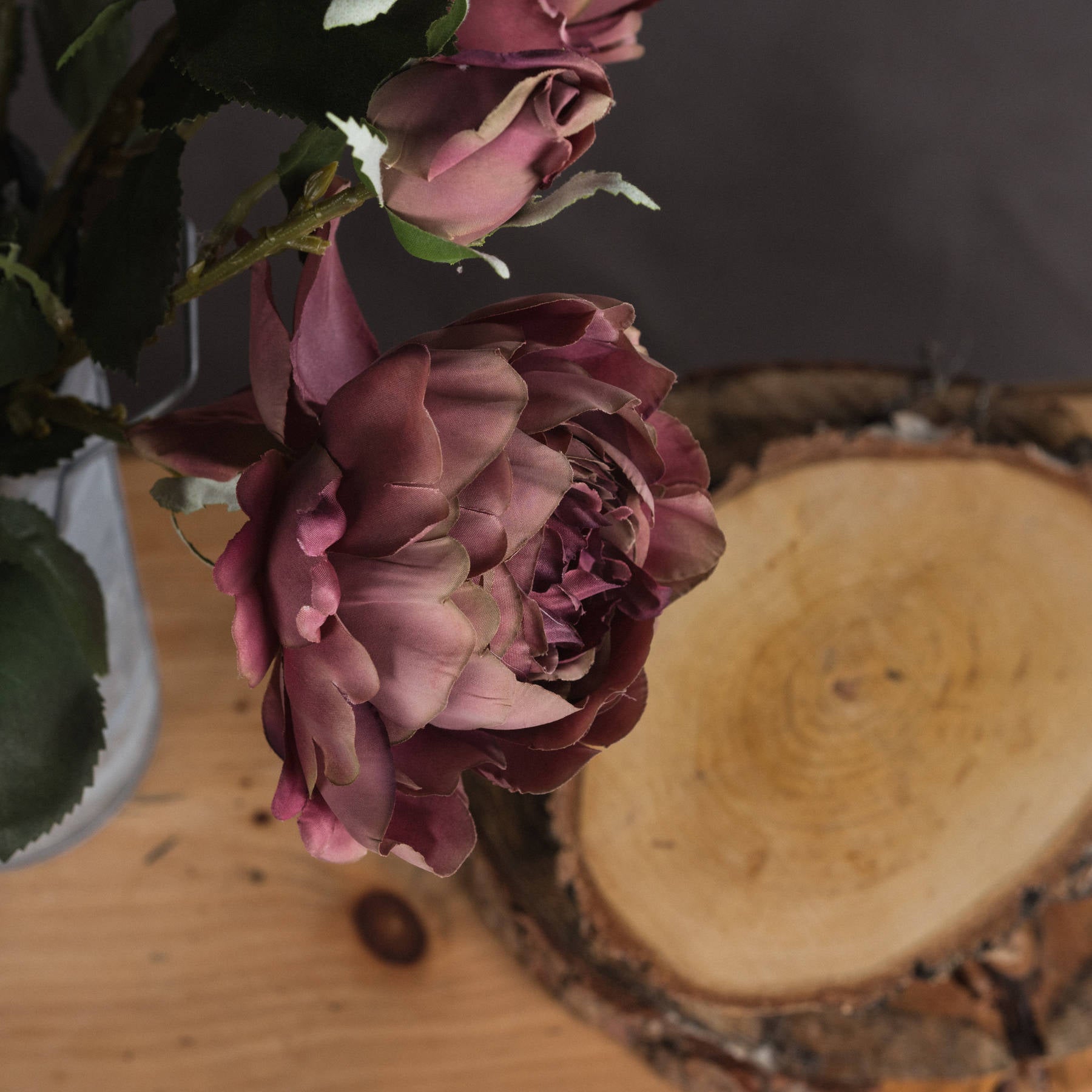 Dusty pink spray rose peony with realistic blooms and closed buds, complemented by lush green leaves, on a wooden surface.