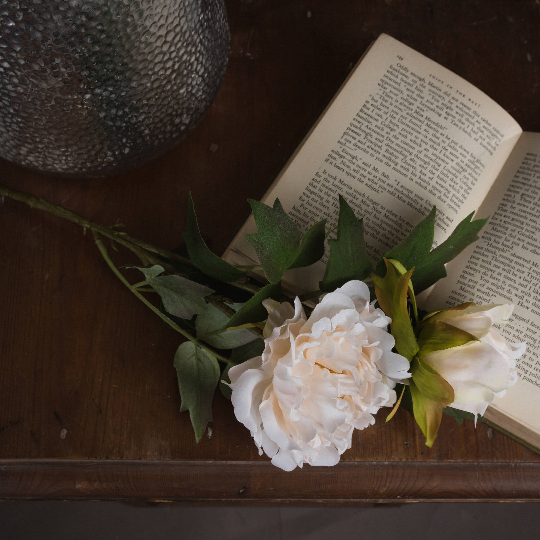 Lifelike white peony with delicate petals and green leaves, resting on a wooden surface beside an open book.