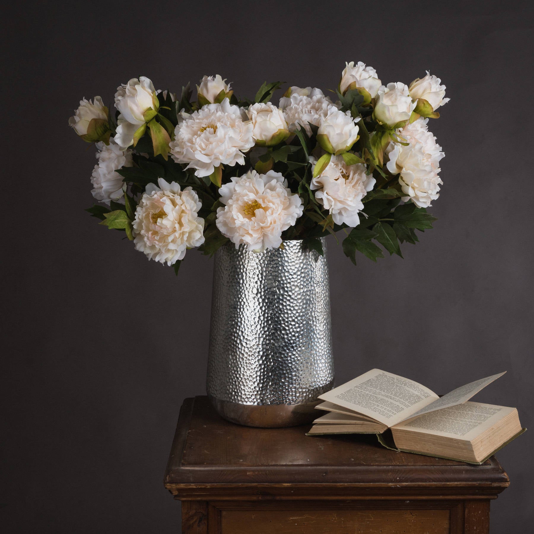 Lifelike white peony bouquet in a hammered silver vase atop a wooden table, accompanied by an open book.