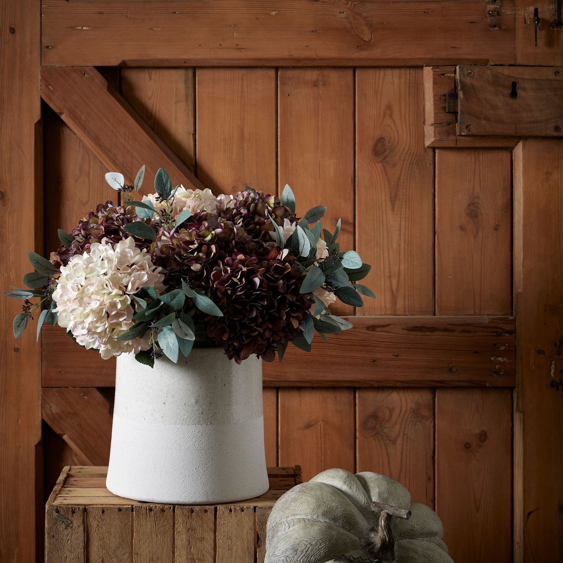 Lifelike hydrangea stem in warm coffee tones, arranged in a textured white pot with soft greenery, set against a wooden backdrop.