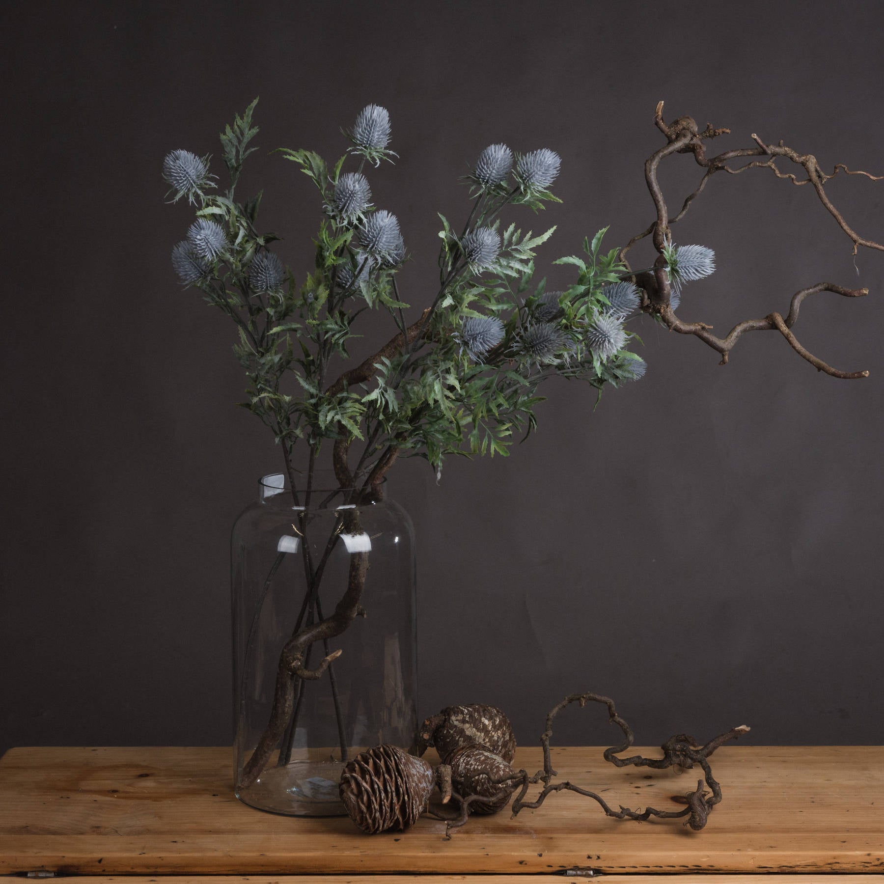 Lifelike blue thistle stems in a clear vase, surrounded by rustic accents on a wooden table, against a dark backdrop.
