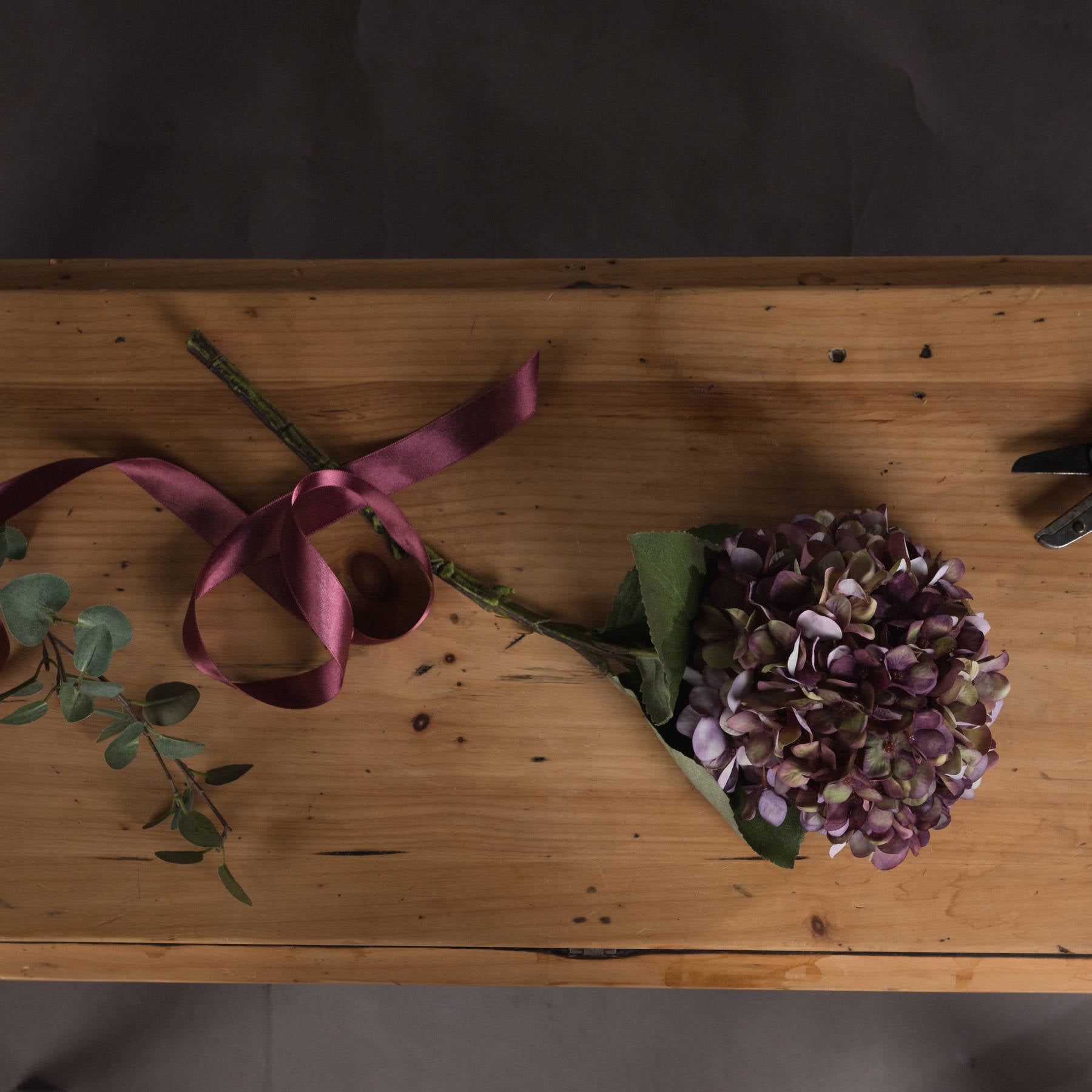 Burgundy hydrangea stem with hand-painted petals in rich purple, green, and burgundy, accompanied by a satin ribbon and leaves on a wooden surface.