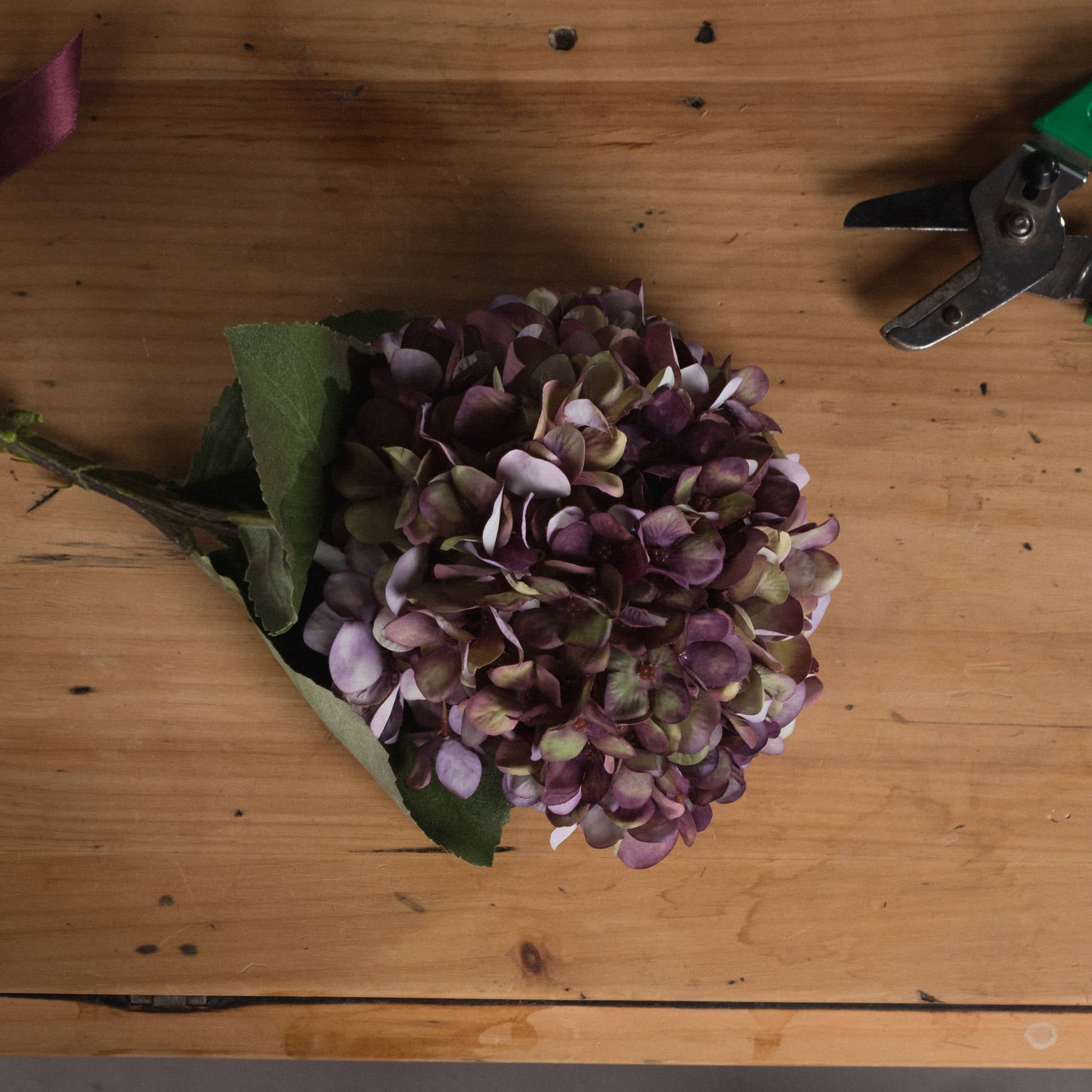 Burgundy hydrangea stem featuring hand-painted petals in rich purple and green hues, set on a wooden surface.