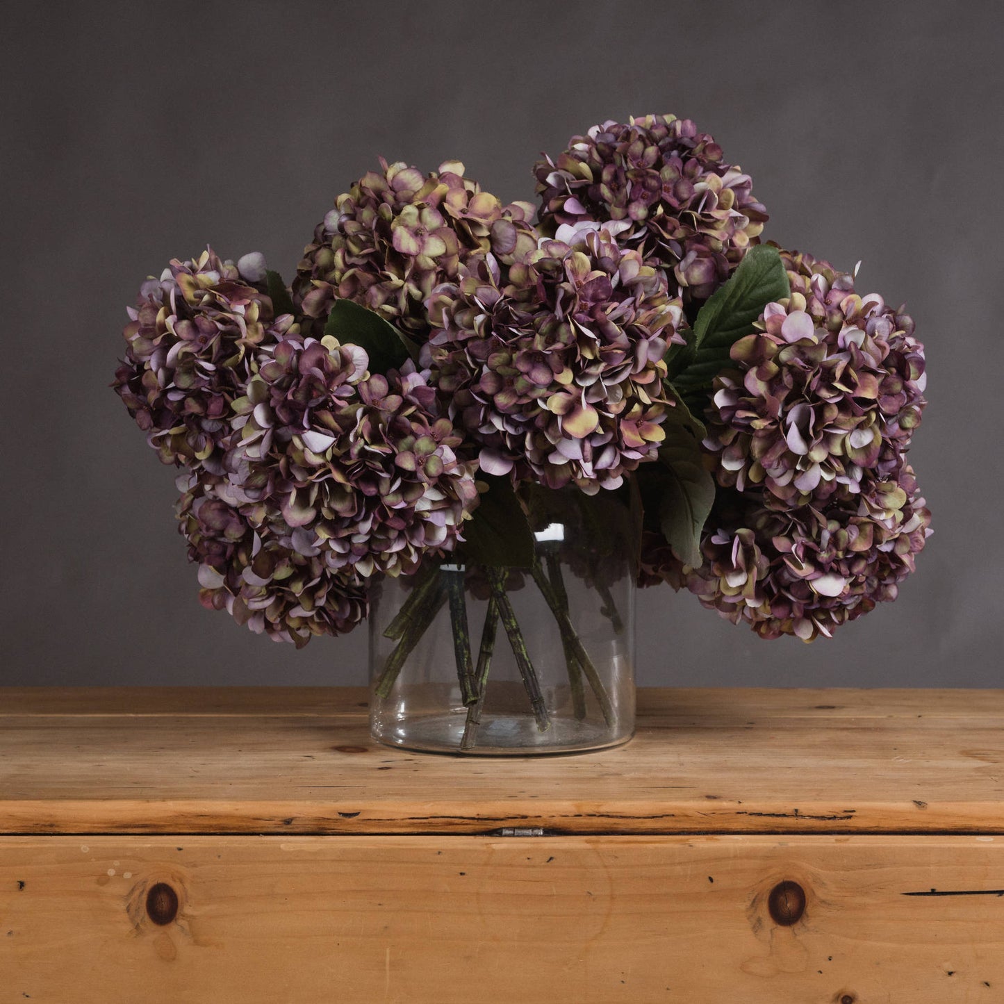 Burgundy hydrangea blooms with rich purple, green, and burgundy hues in a clear vase, set on a wooden surface.