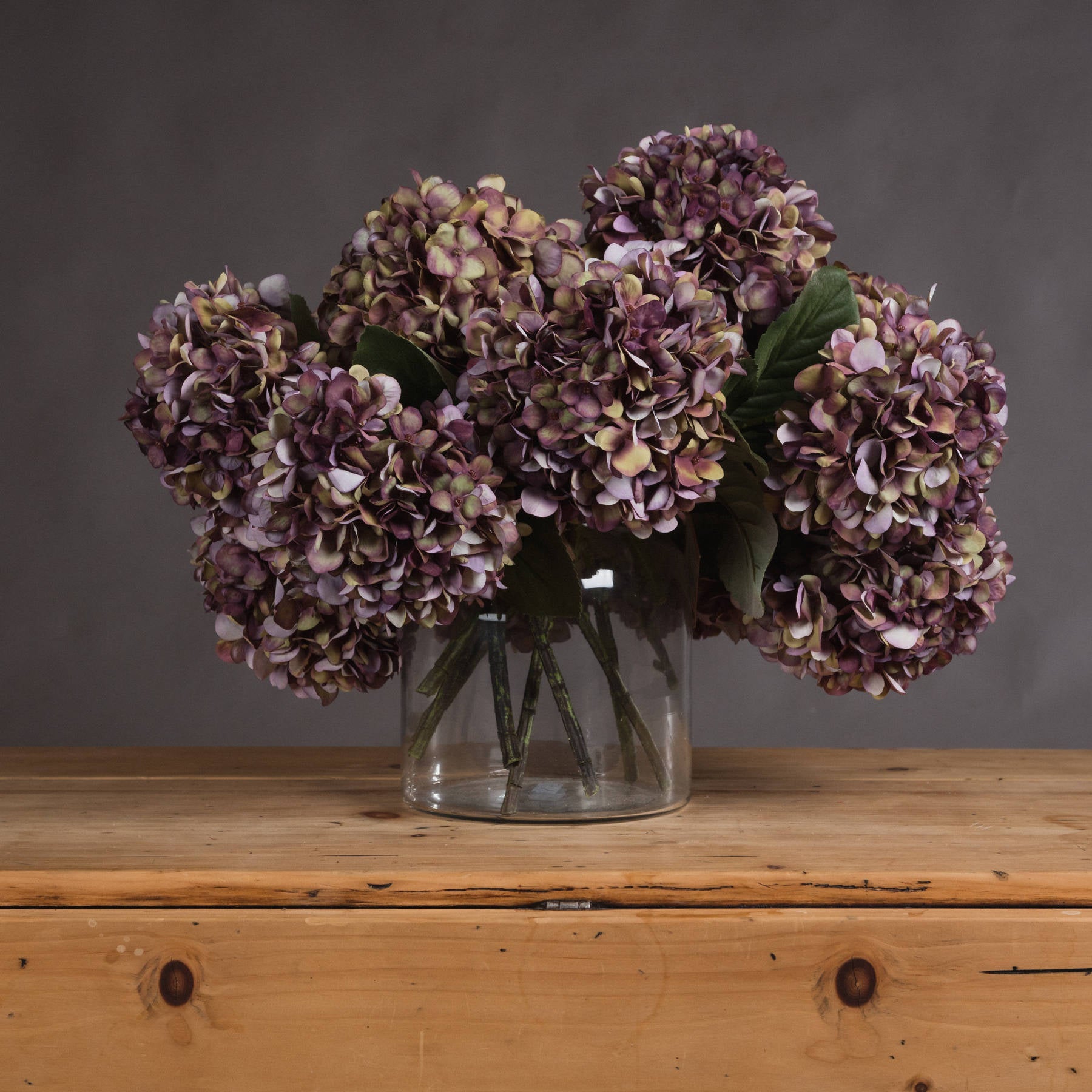 Burgundy hydrangea blooms with rich purple, green, and burgundy hues in a clear vase, set on a wooden surface.