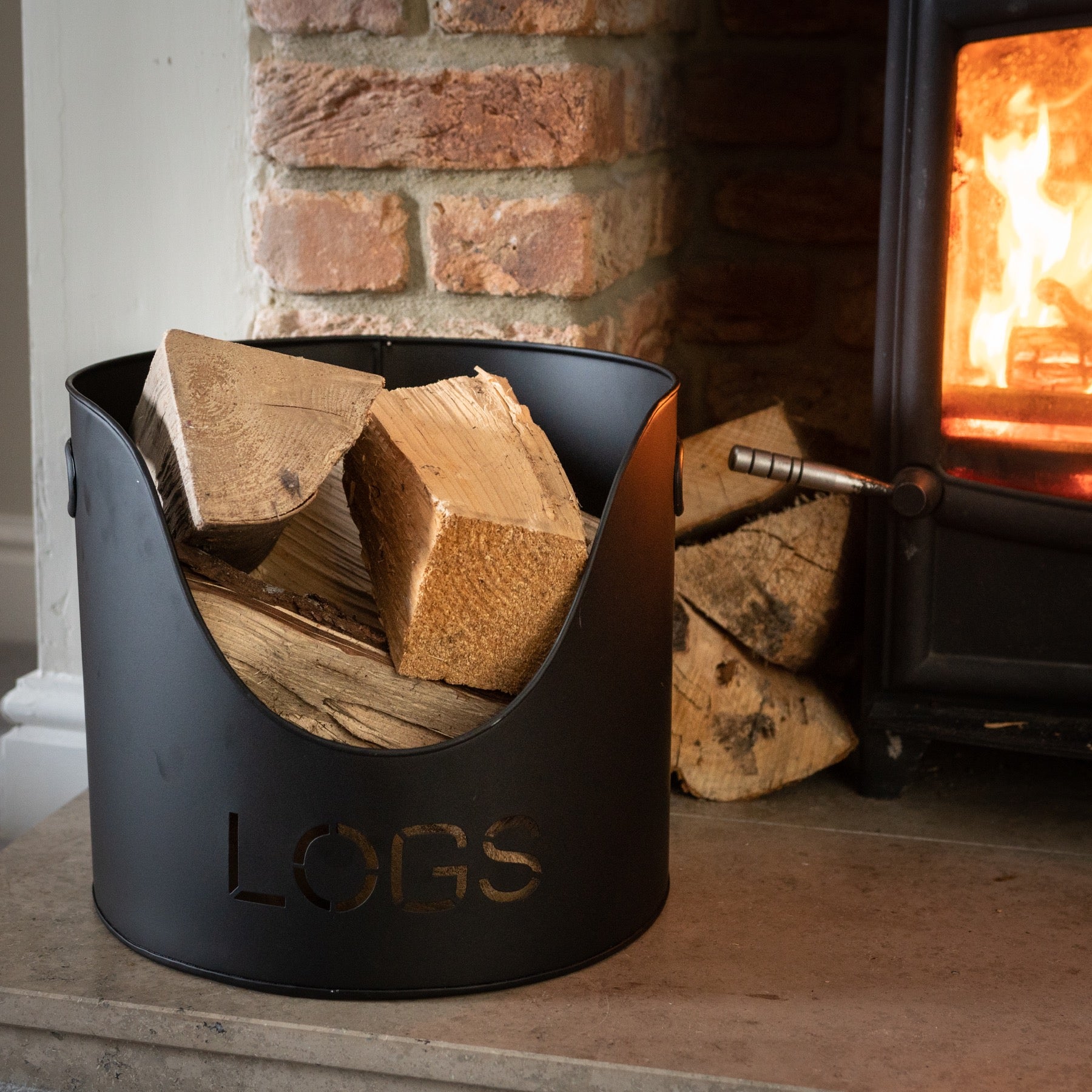 Sleek black bucket for logs, featuring cut-out "LOGS" text and holding neatly stacked firewood beside a cozy fireplace.