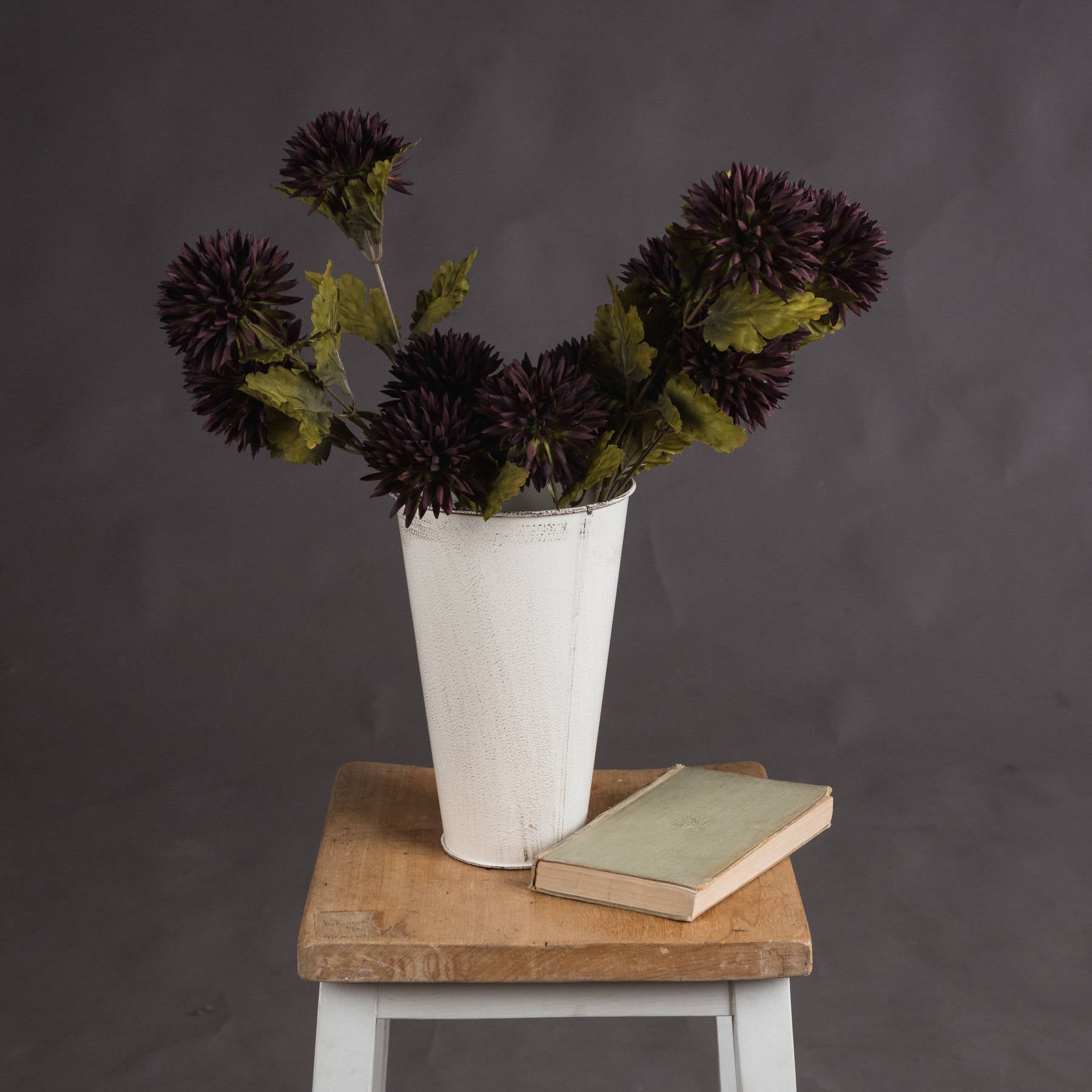 Deep chocolate chrysanthemum stems in a white vase, set on a wooden stool beside a green book, against a gray background.