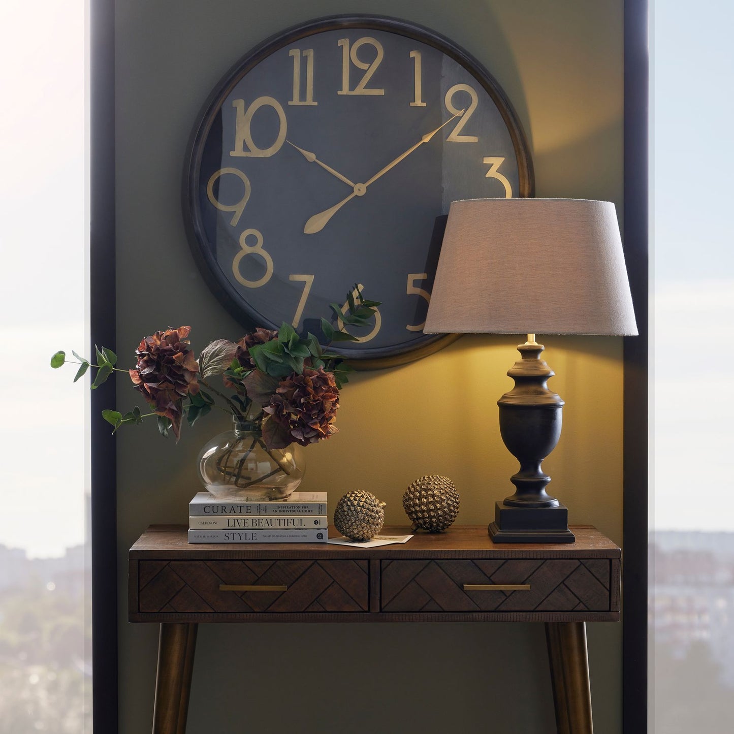 Large black wall clock with brass numerals and hands, exuding vintage charm, above a wooden table with decorative items.
