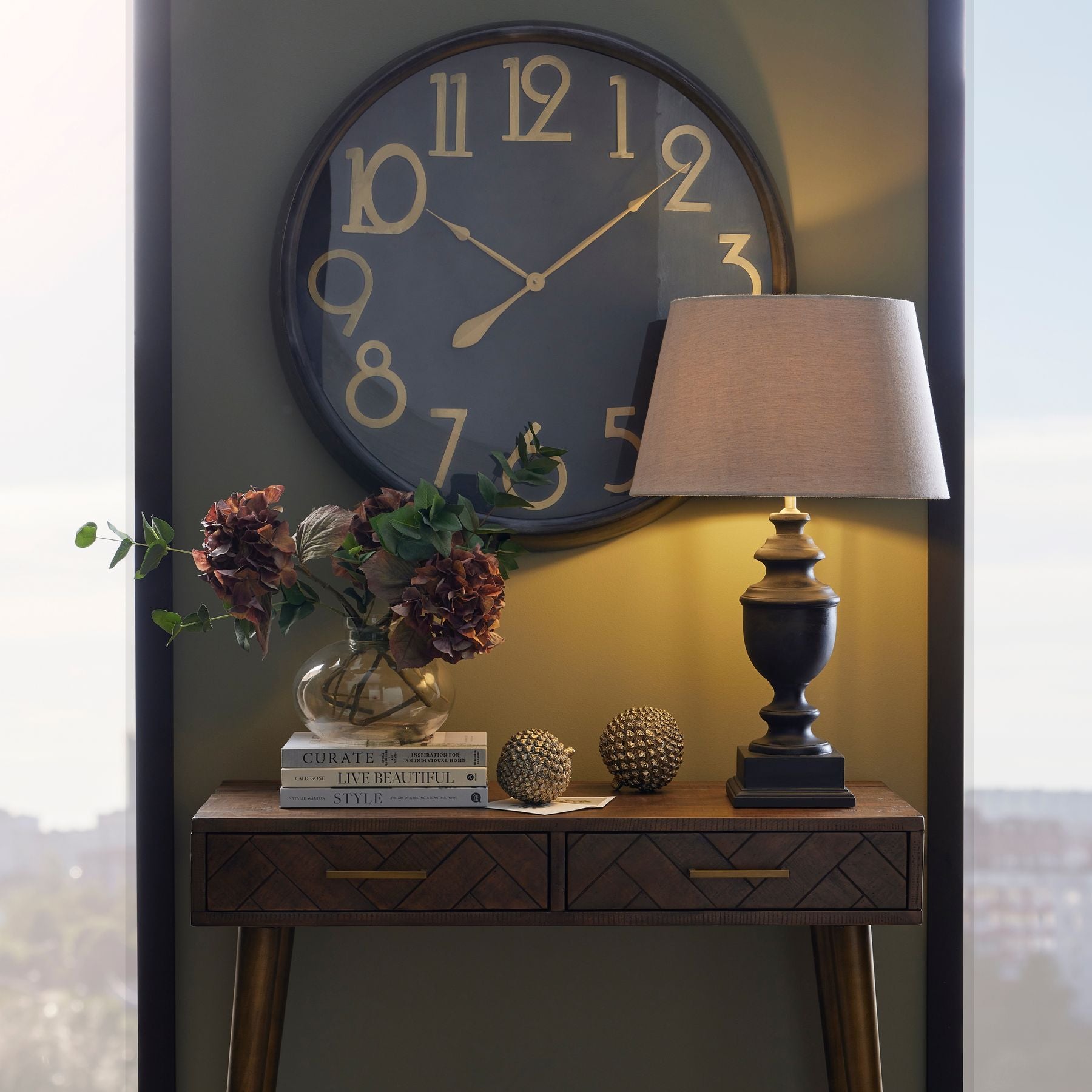 Large black wall clock with brass numerals and hands, exuding vintage charm, above a wooden table with decorative items.