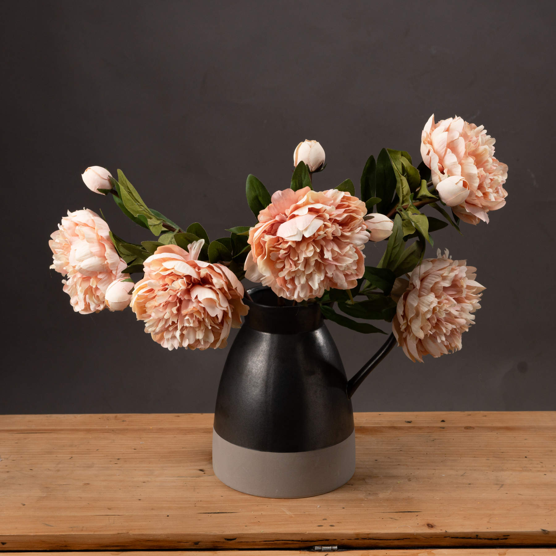 Peach peony roses with realistic petals in soft peach tones, arranged in a modern black and gray pitcher on a wooden surface.