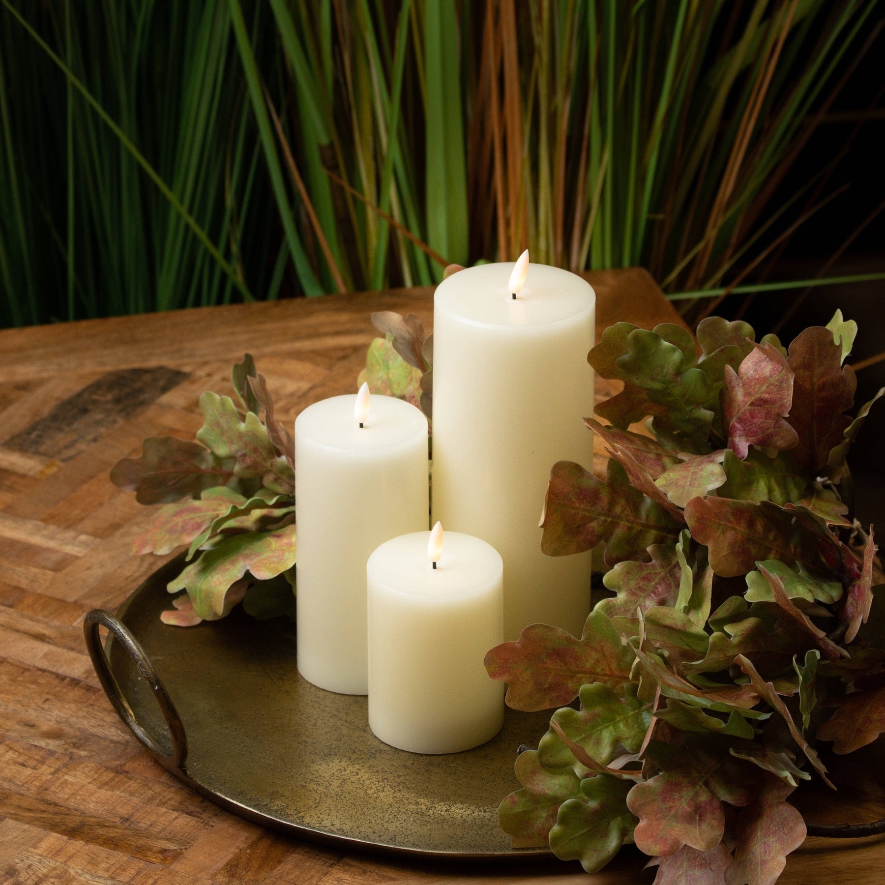 Elegant white LED cream candles in varying heights, set on a brass tray with green and burgundy foliage, enhancing a cozy ambiance.