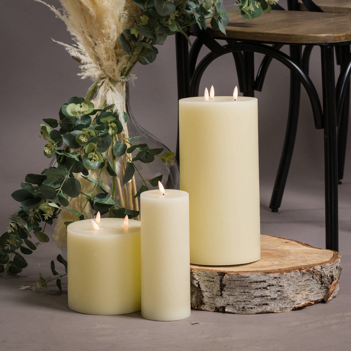Three soft yellow LED cream candles of varying heights, with flickering flames, displayed on a wooden log, surrounded by greenery.