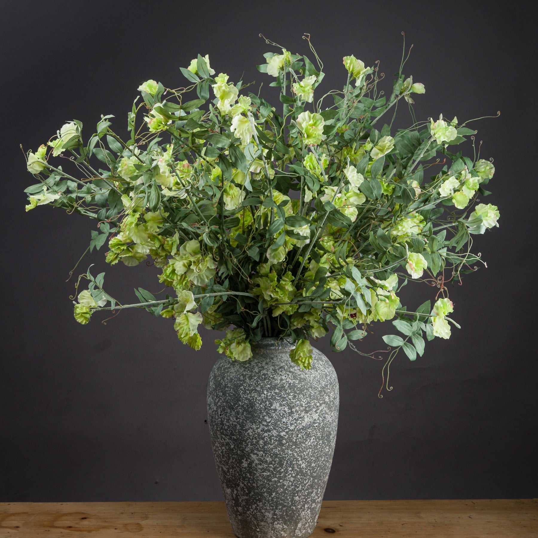Lush artificial Sweet Pea Trail with soft green and cream blossoms, cascading gracefully from a textured gray vase.