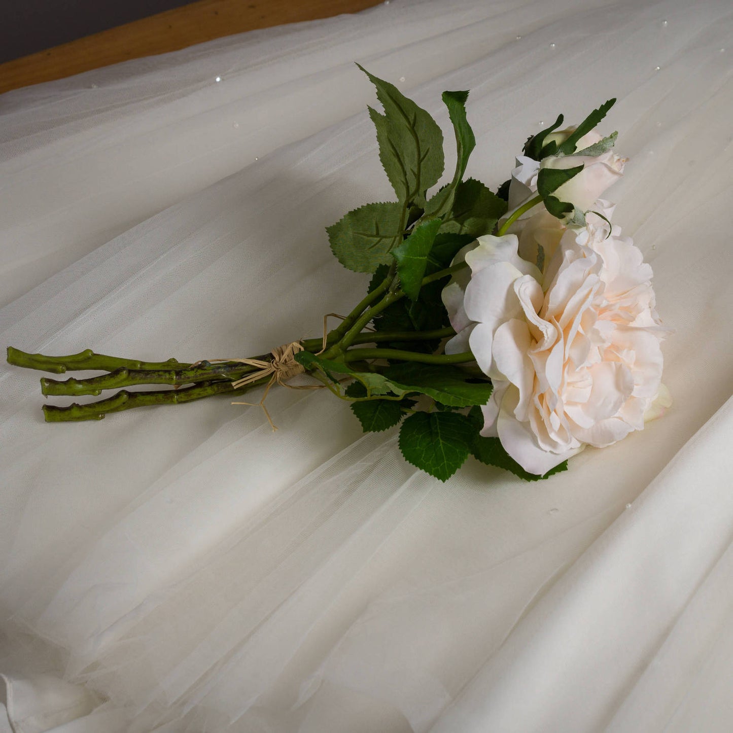 Peachy cream artificial rose bouquet with lush green leaves, bound with rustic twine, resting on soft white fabric.