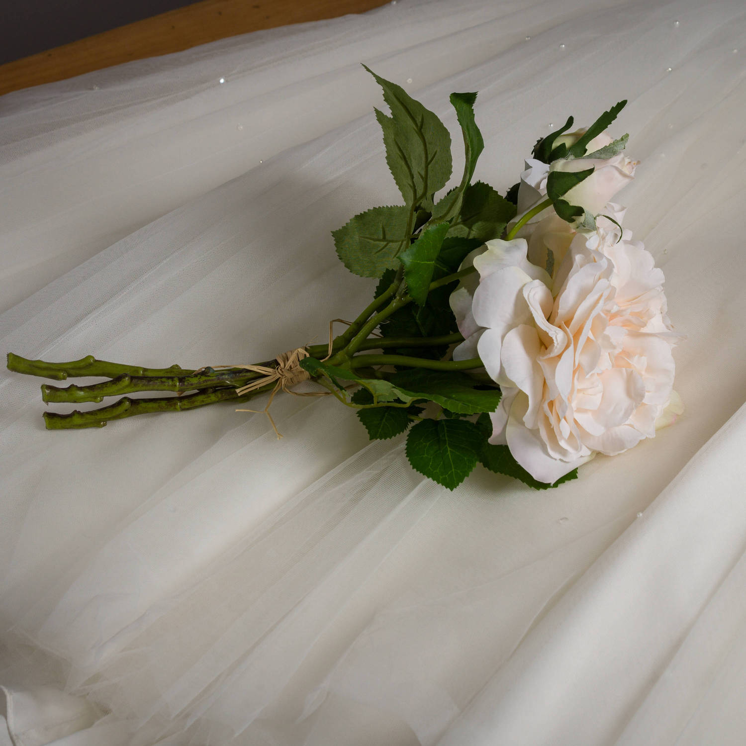 Peachy cream artificial rose bouquet with lush green leaves, bound with rustic twine, resting on soft white fabric.