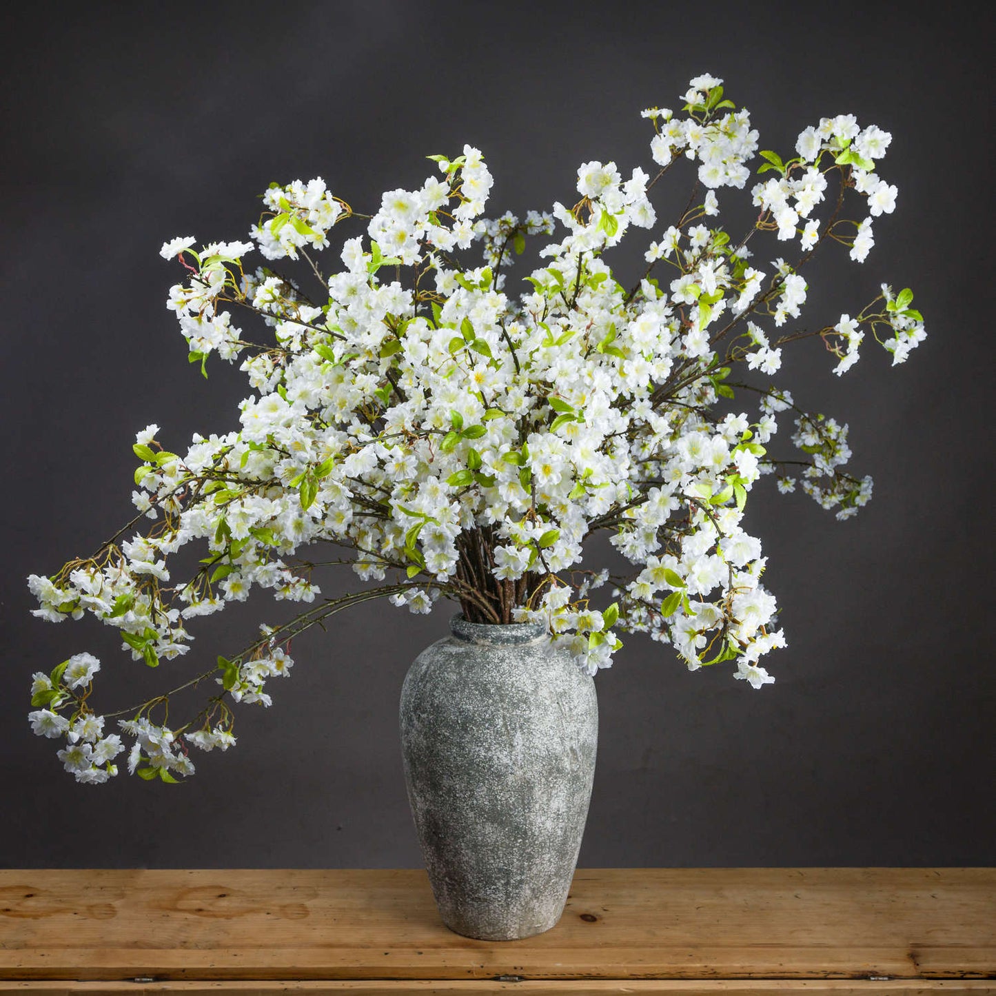 Lifelike white cherry blossom spray in a textured gray vase, showcasing full clusters and delicate green accents, perfect for elegant decor.