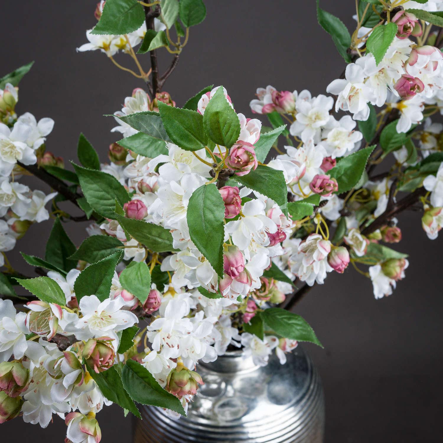 Delicate pale pink and white blossoms with vibrant green leaves on a natural-looking stem, arranged elegantly in a silver vase.