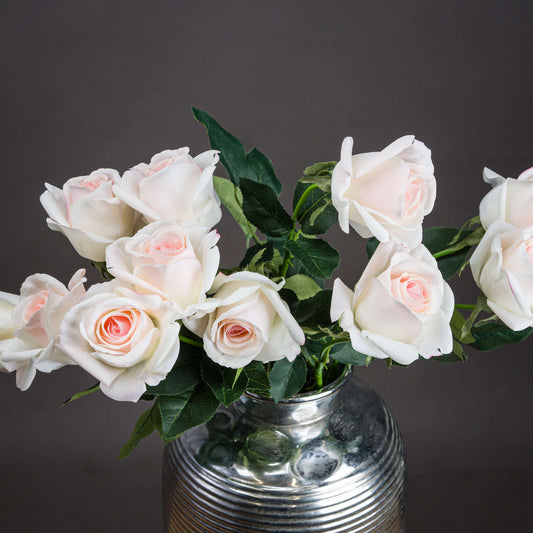 Pale pink artificial tea roses with soft petals and lush green leaves, arranged in a silver vase against a dark background.