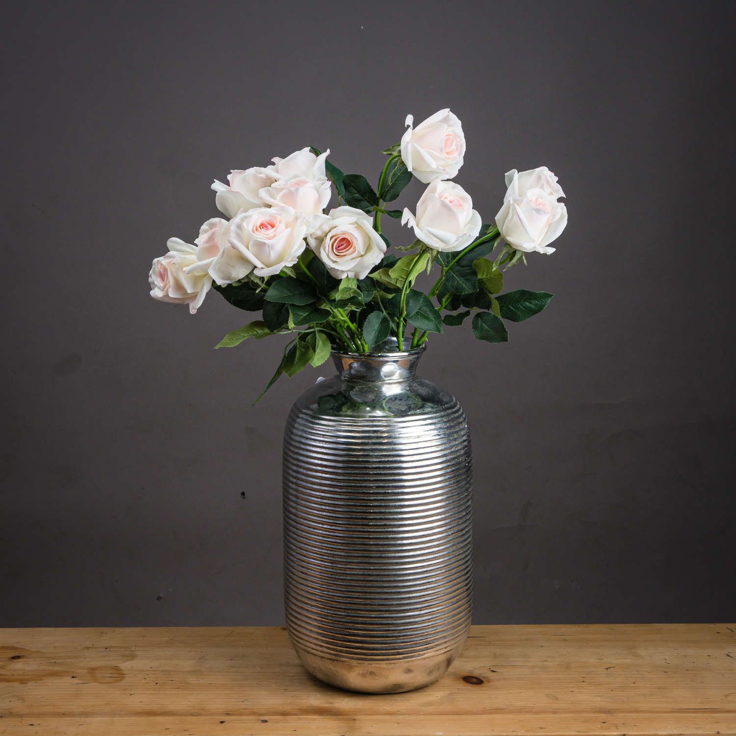 Soft blush pink artificial tea roses bloom gracefully among vibrant green leaves in a sleek, silver-striped vase.