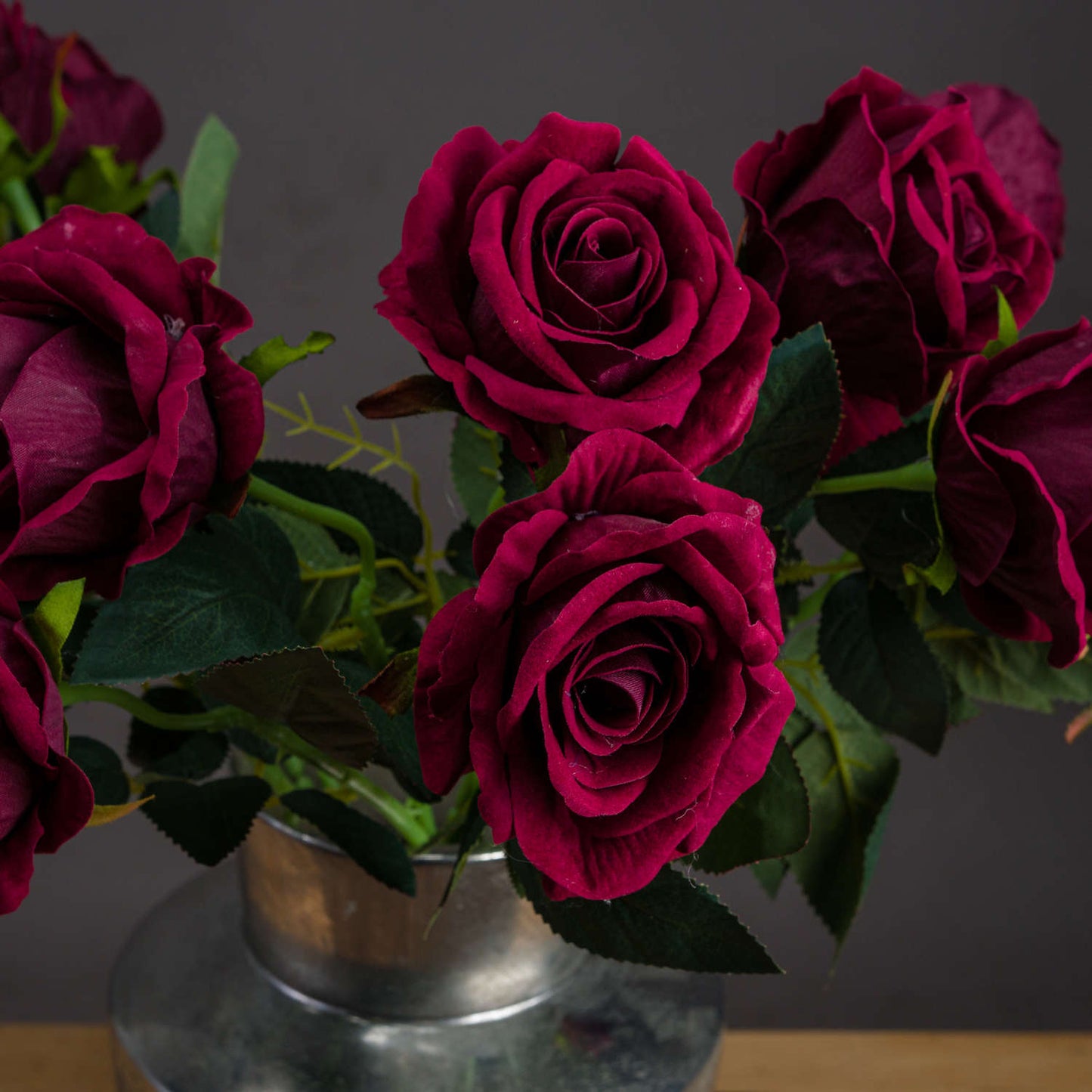 Deep red artificial roses with velvety petals and lush green leaves in a silver vase, exuding elegance and luxury.