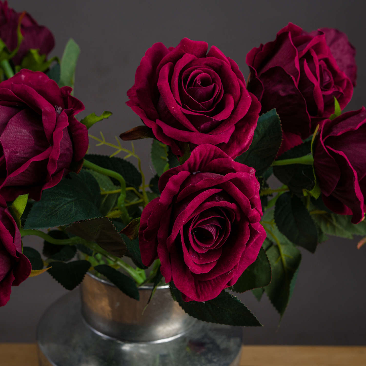 Deep red artificial roses with velvety petals and lush green leaves in a silver vase, exuding elegance and luxury.
